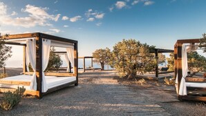A series of wooden bed structures with white curtains, set on a gravel path with trees and a clear sky in the background. - Villas Salobre (San Bartolome de Tirajana)