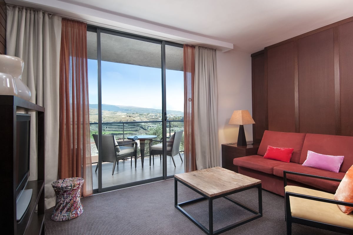 Ein Hotelzimmer mit einer roten Couch, einem Holztisch, einem Fernseher, einem kleinen Nachttisch mit Lampe und einem Balkon mit Tisch und Stühlen.