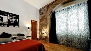A bedroom with a brick wall, a bed with a red blanket, patterned curtains, and a wall-mounted light.