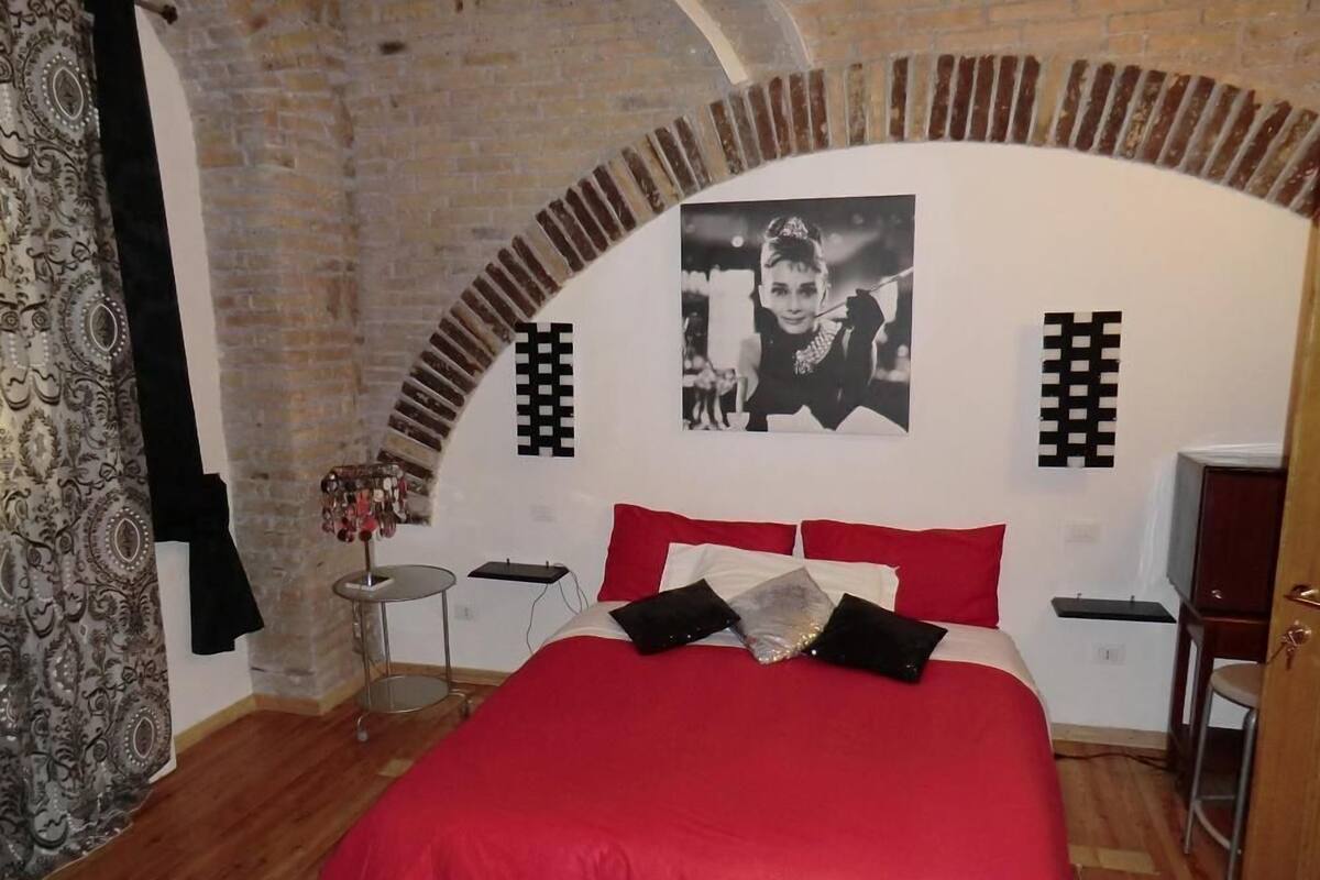 A bedroom with a red bed, a black and white photo on the wall, and a brick archway.