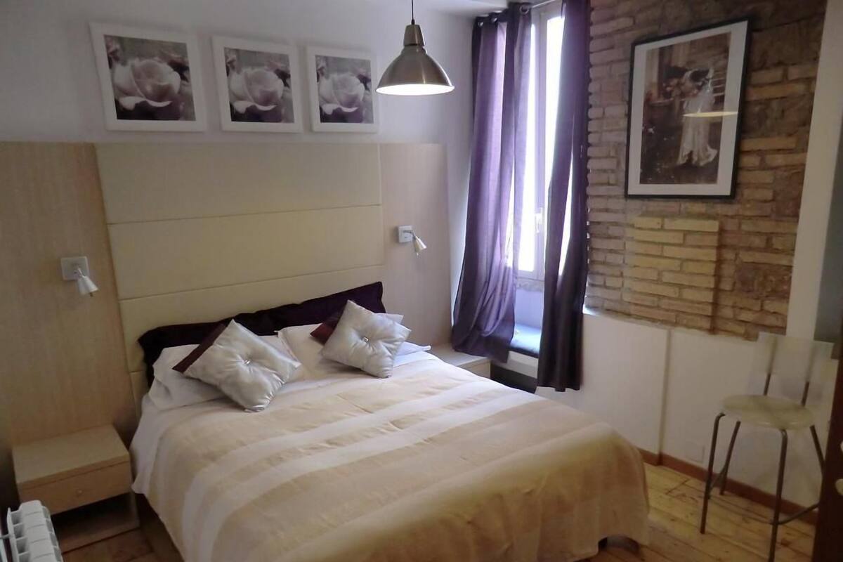 A bedroom with a bed, a headboard, a bedside table, a radiator, a window with curtains, and a wall-mounted picture.