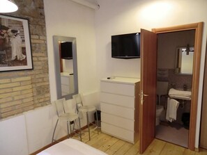 A bedroom with a bed, a TV, a mirror, a dresser, and a bathroom visible through an open door. - Caesar's Rooms (Rome)