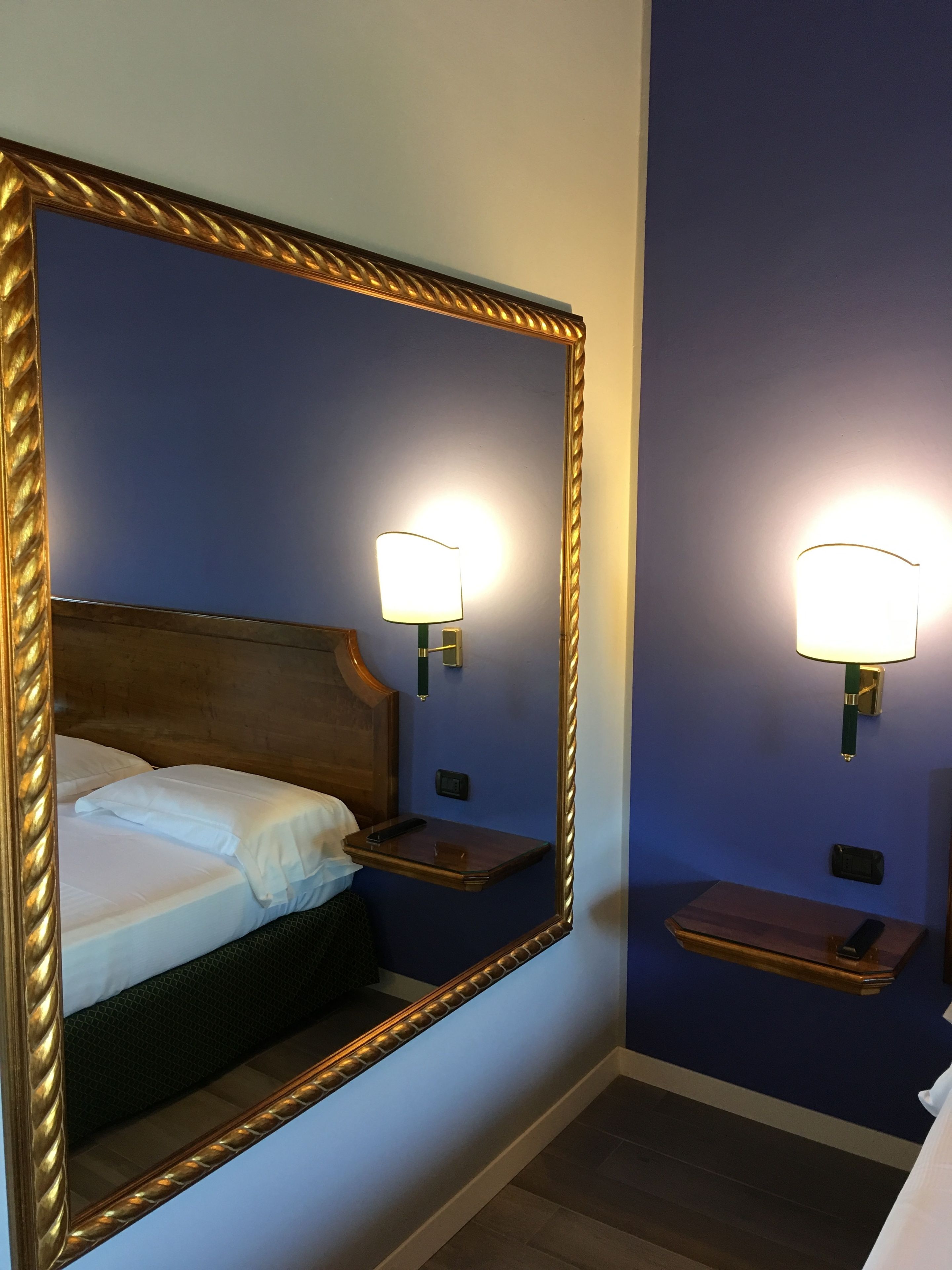Habitación de hotel con un espejo grande, una cama con ropa blanca, dos mesitas de noche con lámparas y un cabecero de madera.