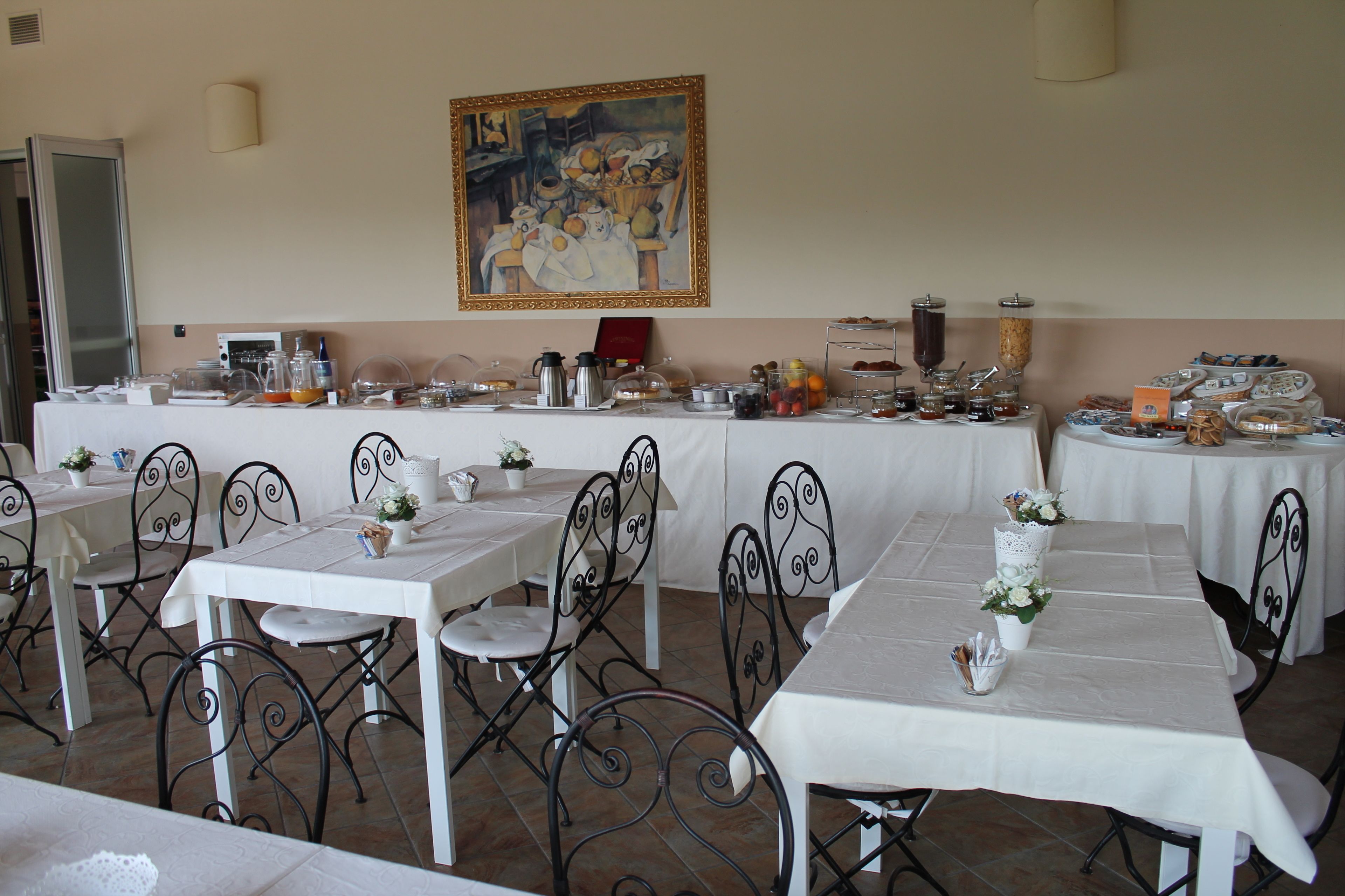 A dining area with white tables and black metal chairs, a painting on the wall, and various food and drink items on the tables.