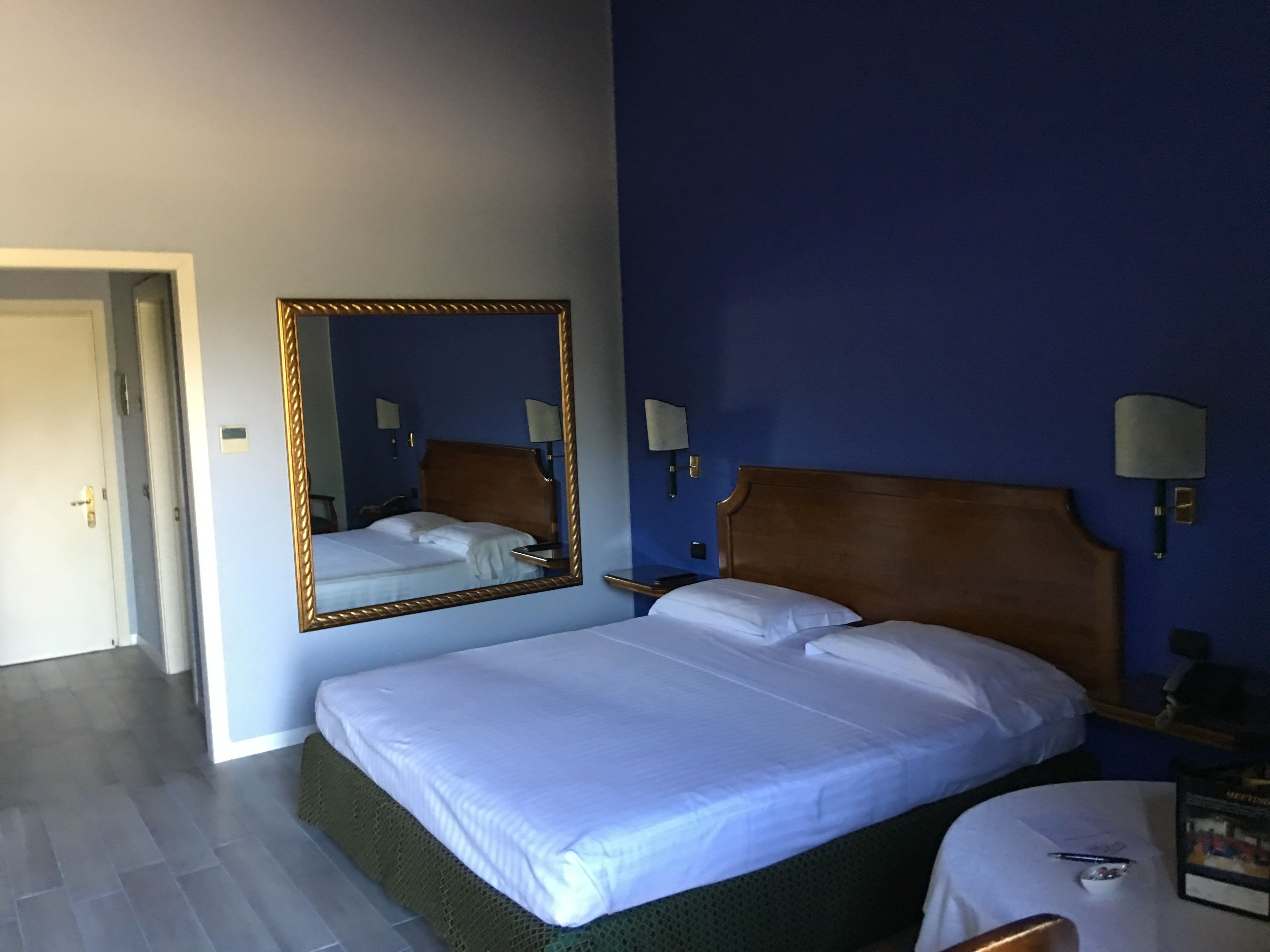 A hotel room with a large bed, a mirror, two bedside lamps, and a small table with a pen and a book.