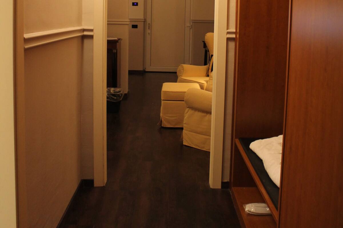 A narrow hallway with a wooden wardrobe on the right, a beige armchair in the center, and a white door at the end.