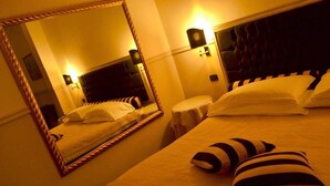 A hotel room with a large mirror, a bed with striped pillows, a round bedside table, and a headboard with a tufted design. - Hotel Don Carlo (Broni)