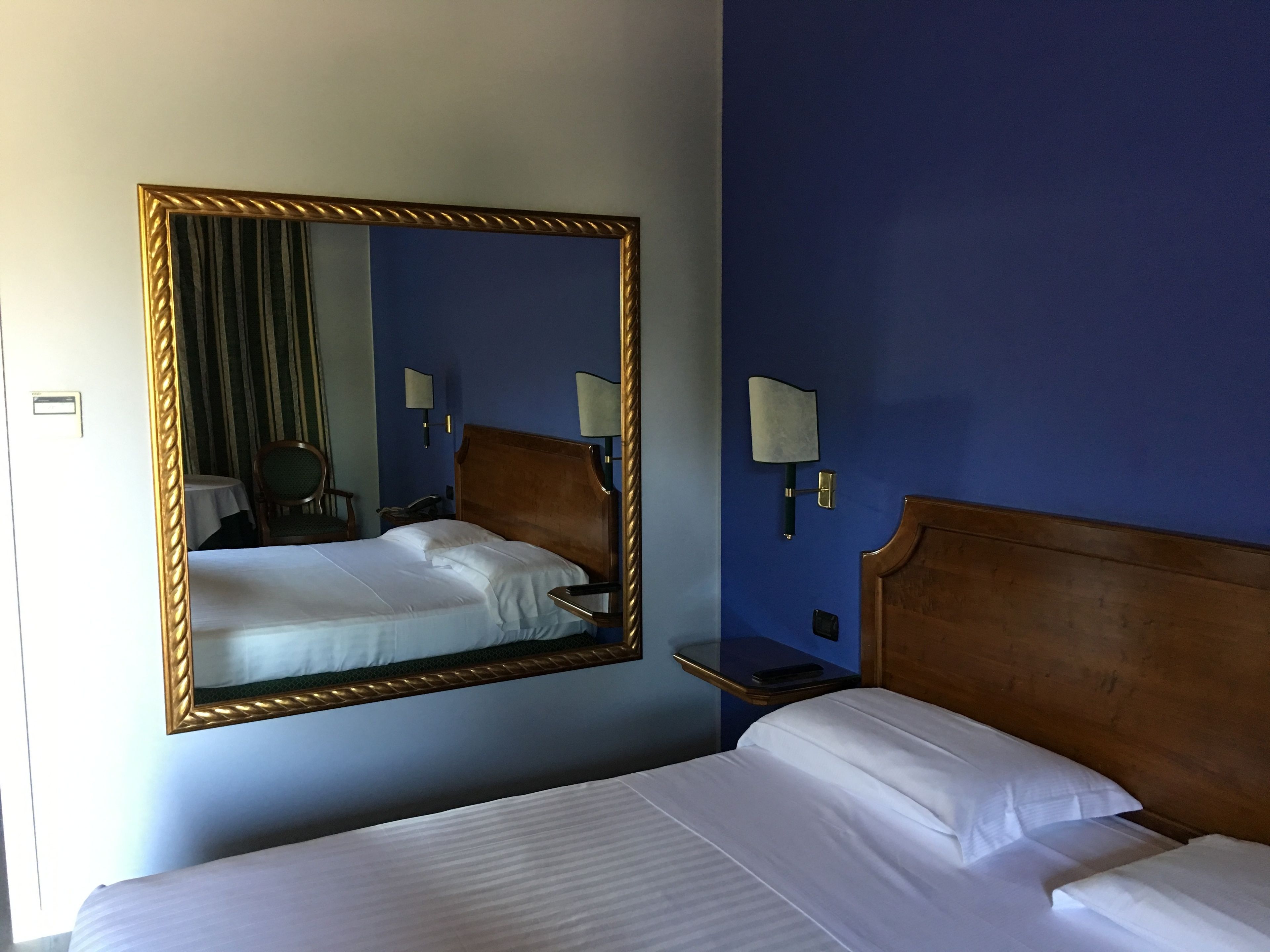 Una habitación de hotel con un espejo grande, un cabecero de madera, dos camas con sábanas blancas, una mesita de noche y dos lámparas fijadas a la pared.
