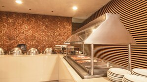 Uma área de buffet com uma parede de padrão geométrico, um expositor de alimentos em aço inoxidável e uma pilha de pratos brancos.