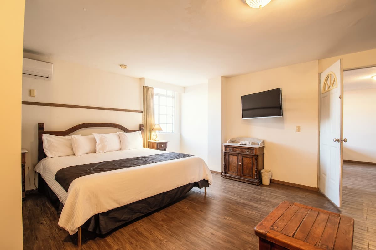 Un dormitorio con cama, piso de madera, una mesita de noche, lámpara, televisor montado en la pared y una puerta.