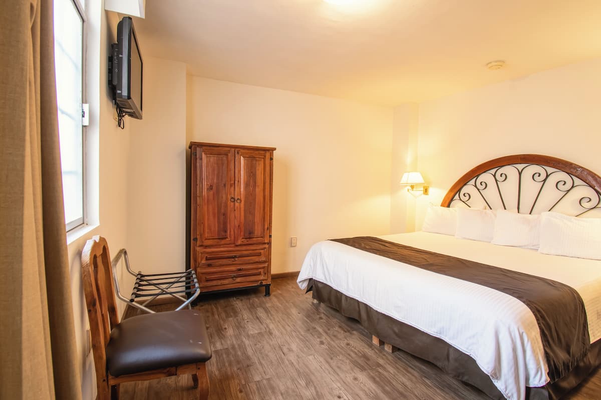 Un dormitorio con un armario de madera, una cama con ropa de cama blanca y oscura, una silla y una lámpara en la pared.