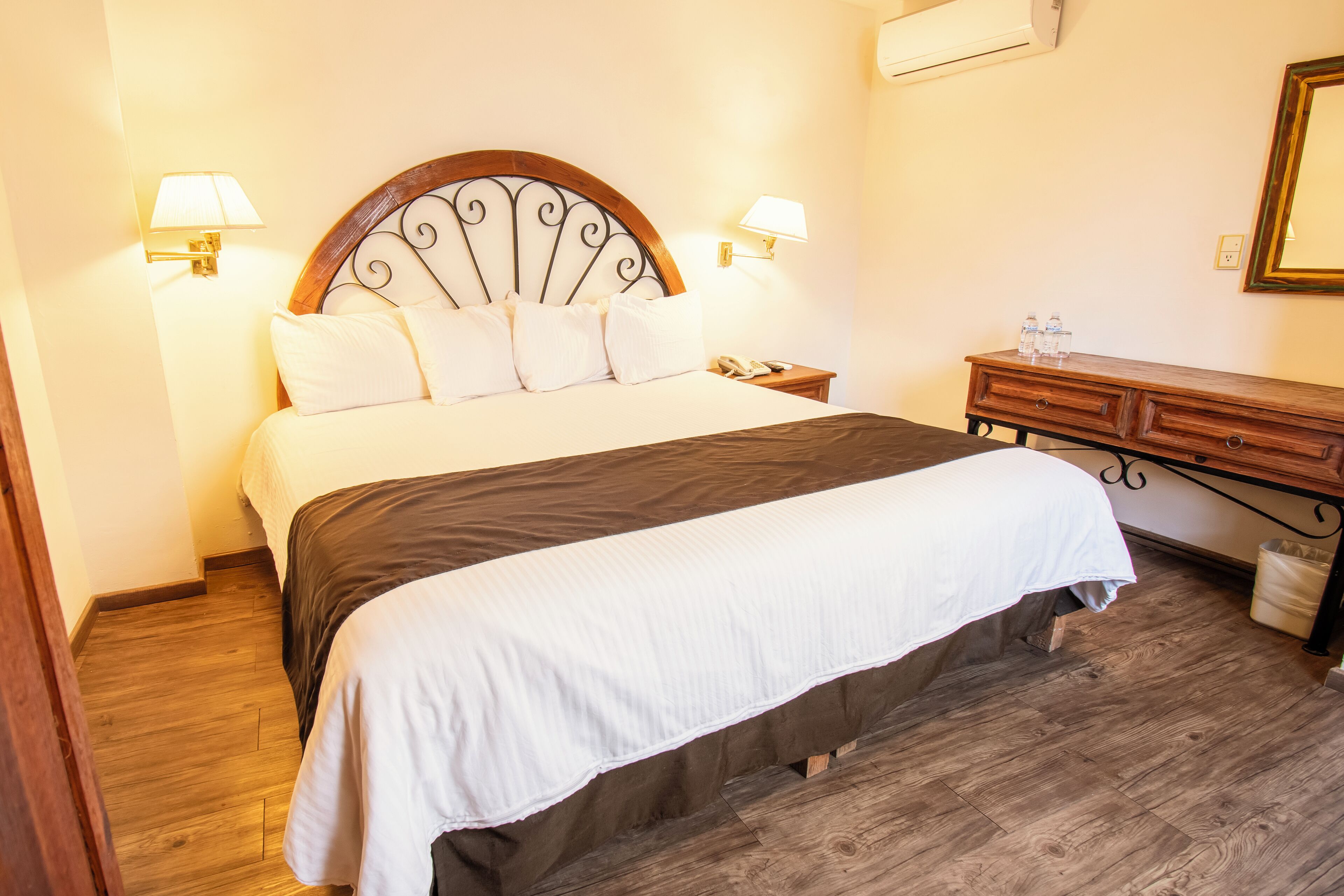 A neatly made bed with a wooden headboard, a bedside table with a lamp, and a wooden dresser with a mirror.