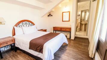 Une chambre à coucher avec un lit en bois, une table de chevet, une commode et une salle de bain visible à travers une porte ouverte.