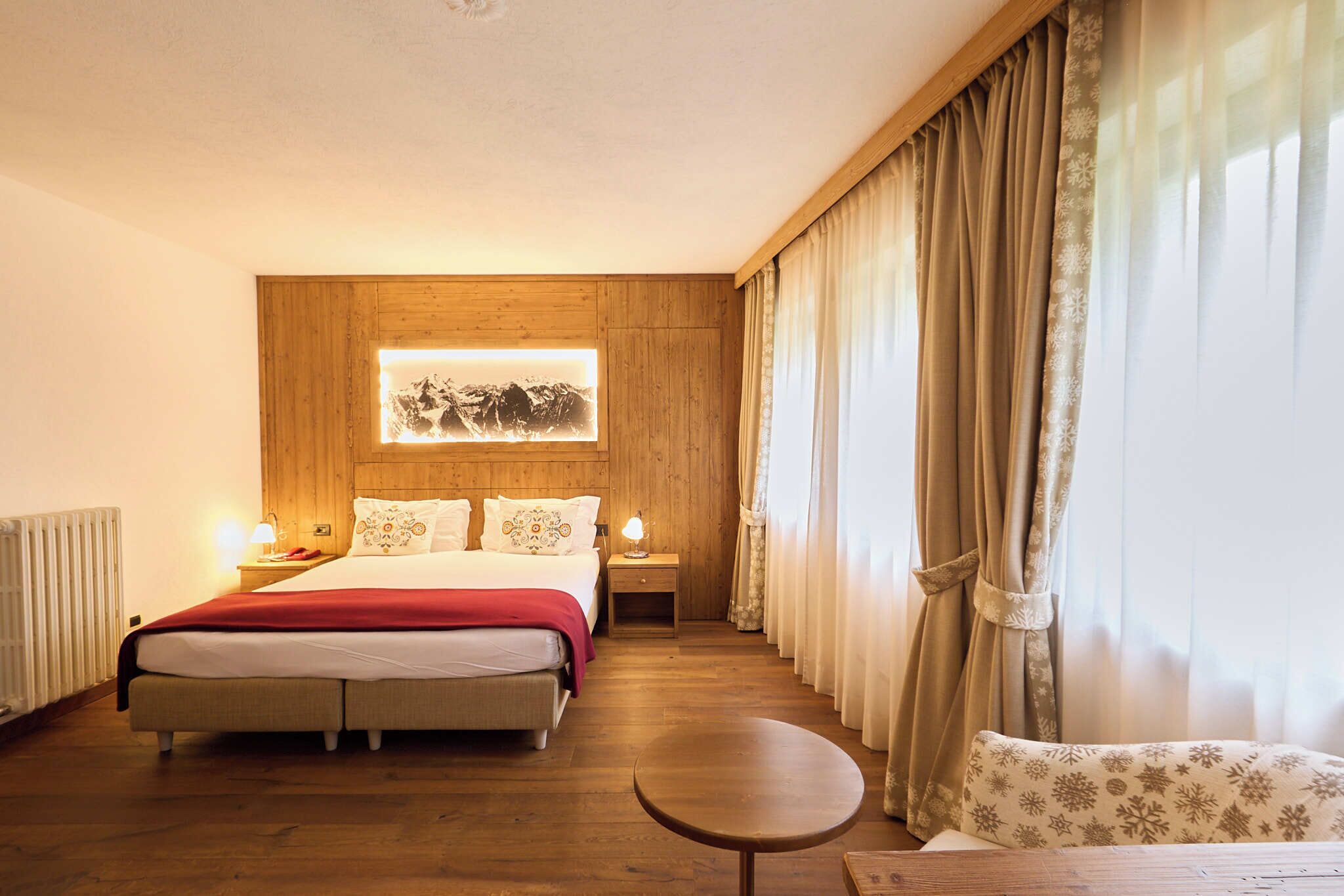 Ein Hotelzimmer mit hölzernem Kopfteil, einem Bett mit roter Decke, einem Nachttisch mit Lampe, einem runden Tisch und Blick auf einen Berg durch ein fensterbelesertes Fenster.