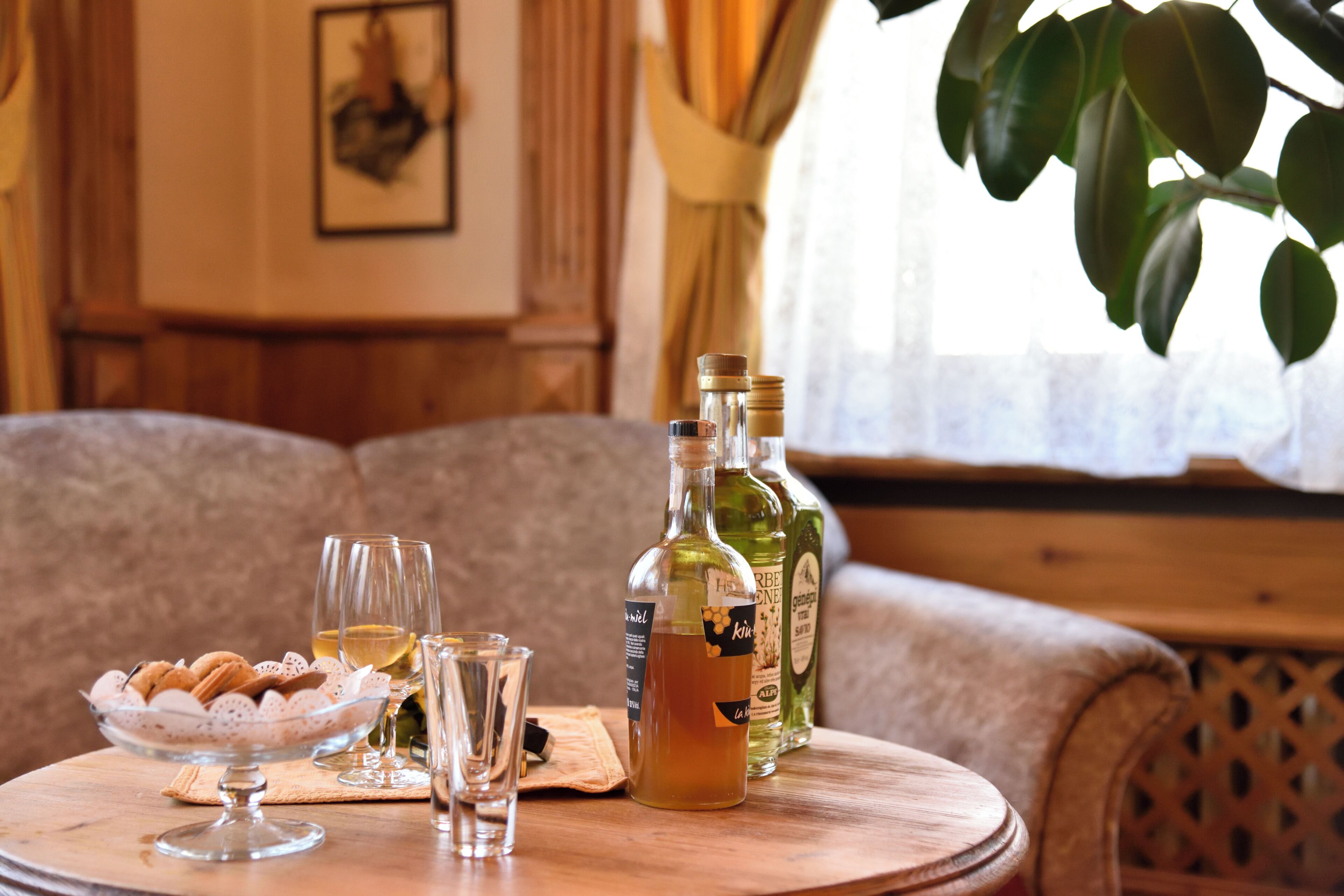 A round wooden table with a bowl of cookies, two glasses, and three bottles of liquor.