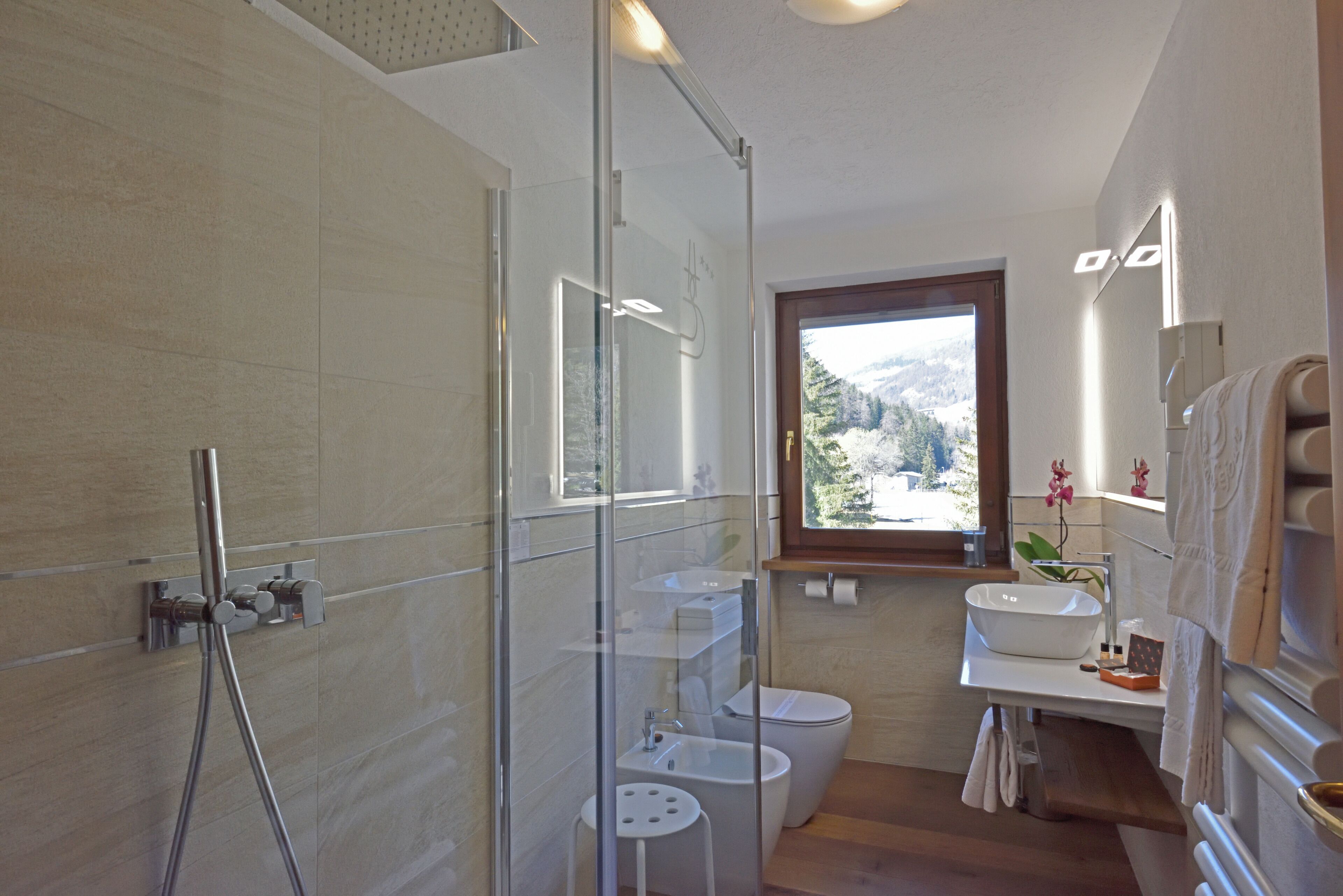 Et moderne badeværelse med brusekabine i glas, toilet, håndvask og et vindue med udsigt over et bjergrigt landskab.