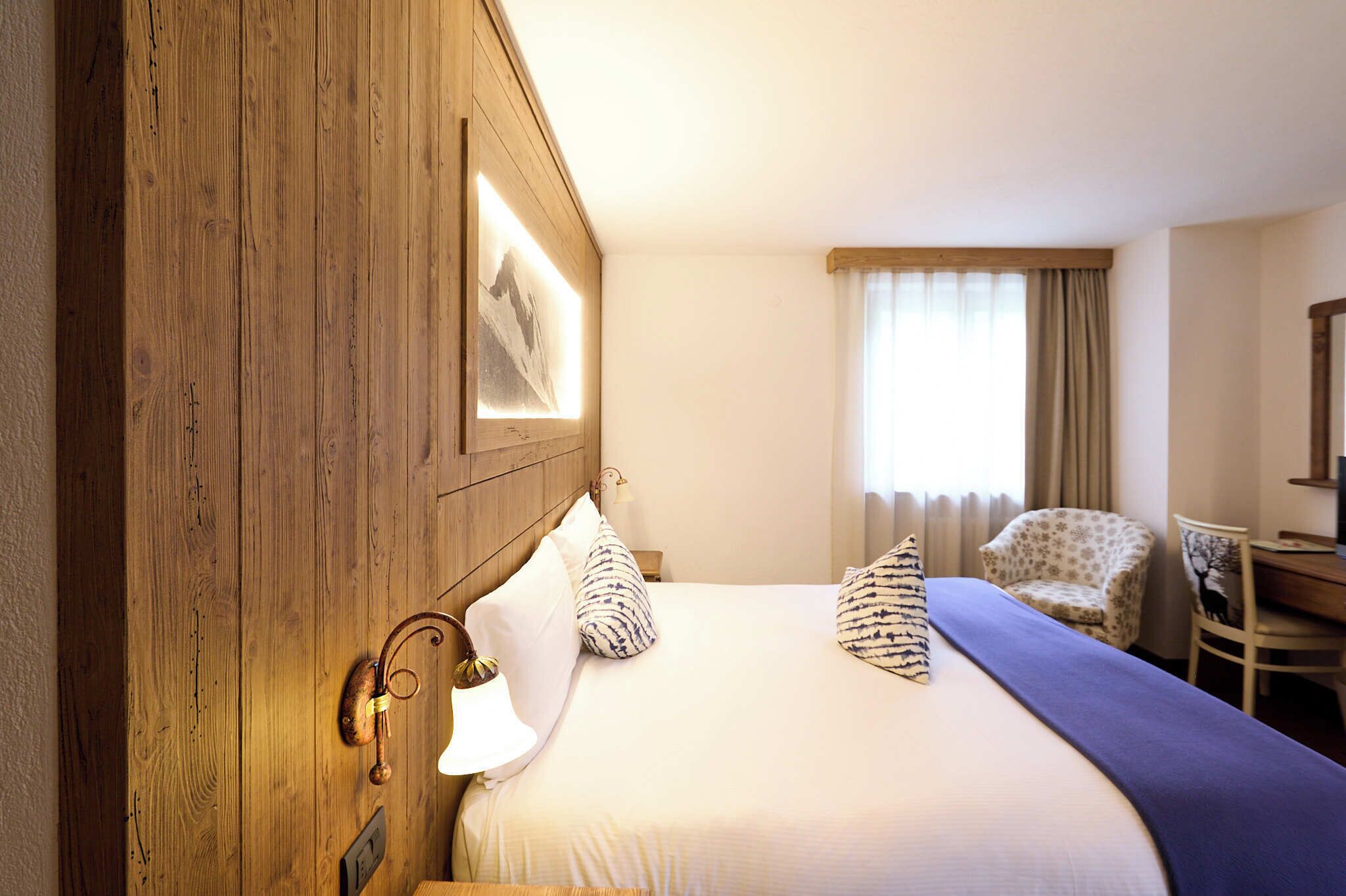 A hotel room with a wooden headboard, a bed with white bedding, a chair, a desk, and a window with curtains.
