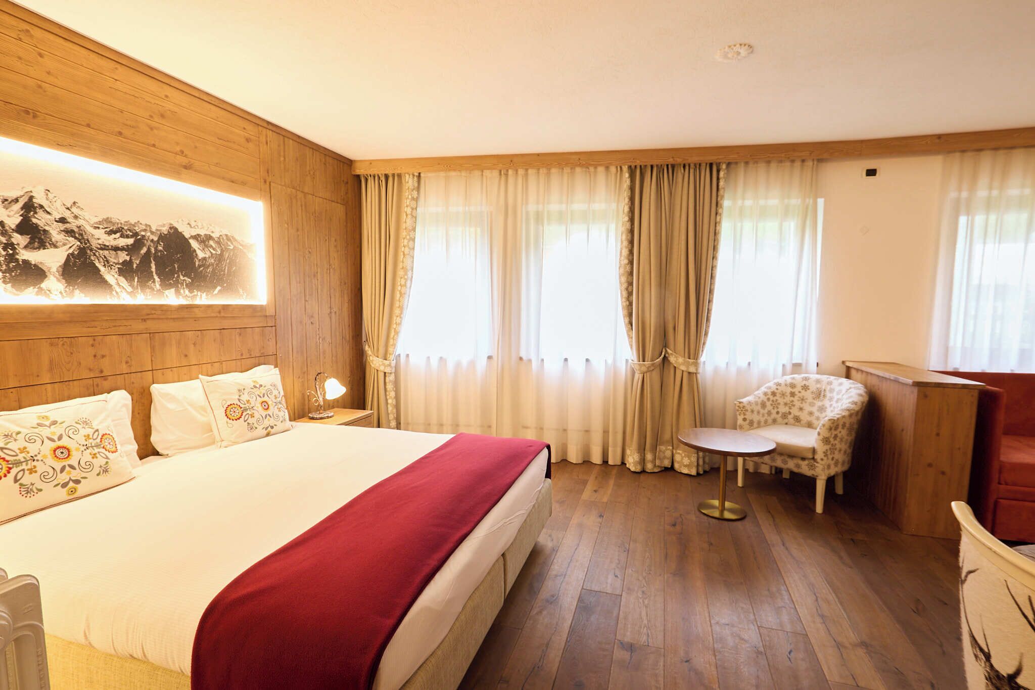 A hotel room with a wooden headboard, a large bed with a red blanket, a small round table, a floral armchair, and a wooden desk.