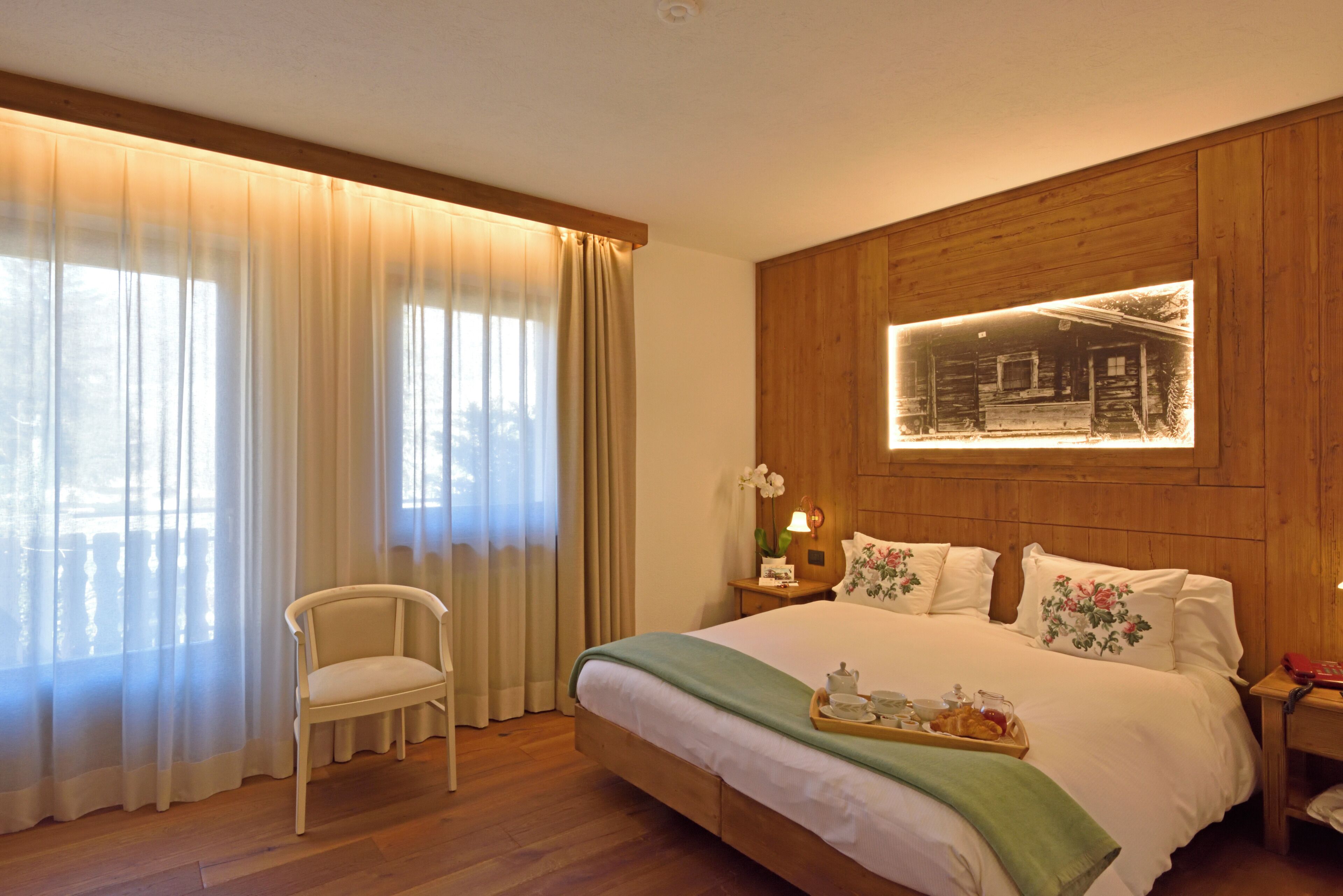 Habitació d'hotel amb un capçal de llit de fusta, un llit amb almofadines florals, una cadira i una nitòria amb una làmpada.