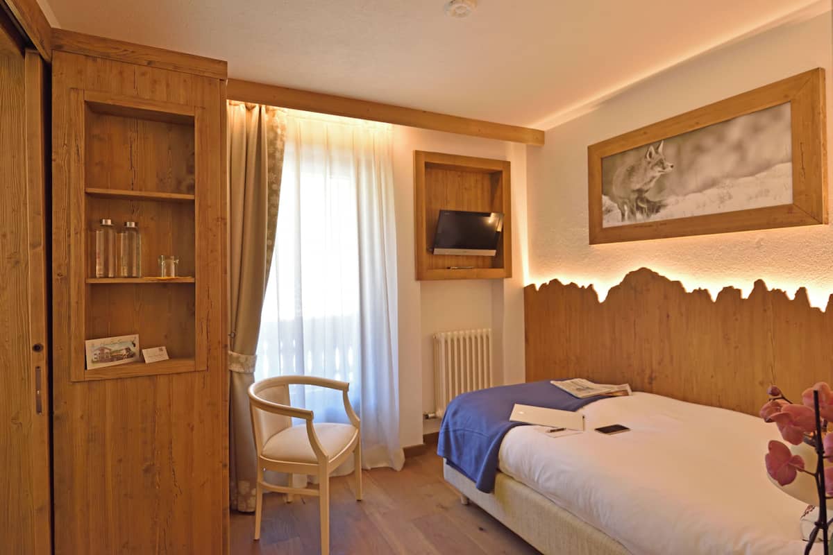 A wooden-framed room with a bed, a chair, a shelf with items, a window with curtains, and a framed picture on the wall.
