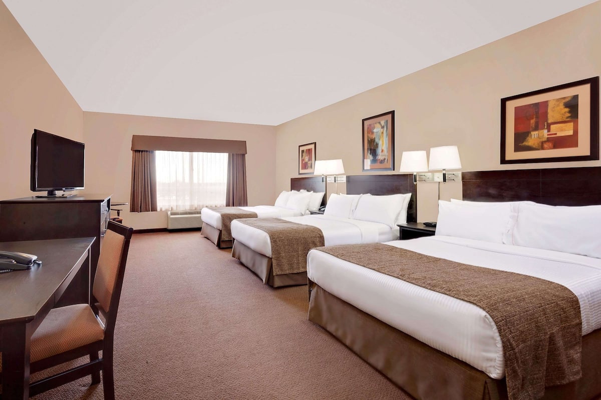 Habitación de hotel con tres camas, un escritorio con silla, televisor y obras de arte en las paredes.
