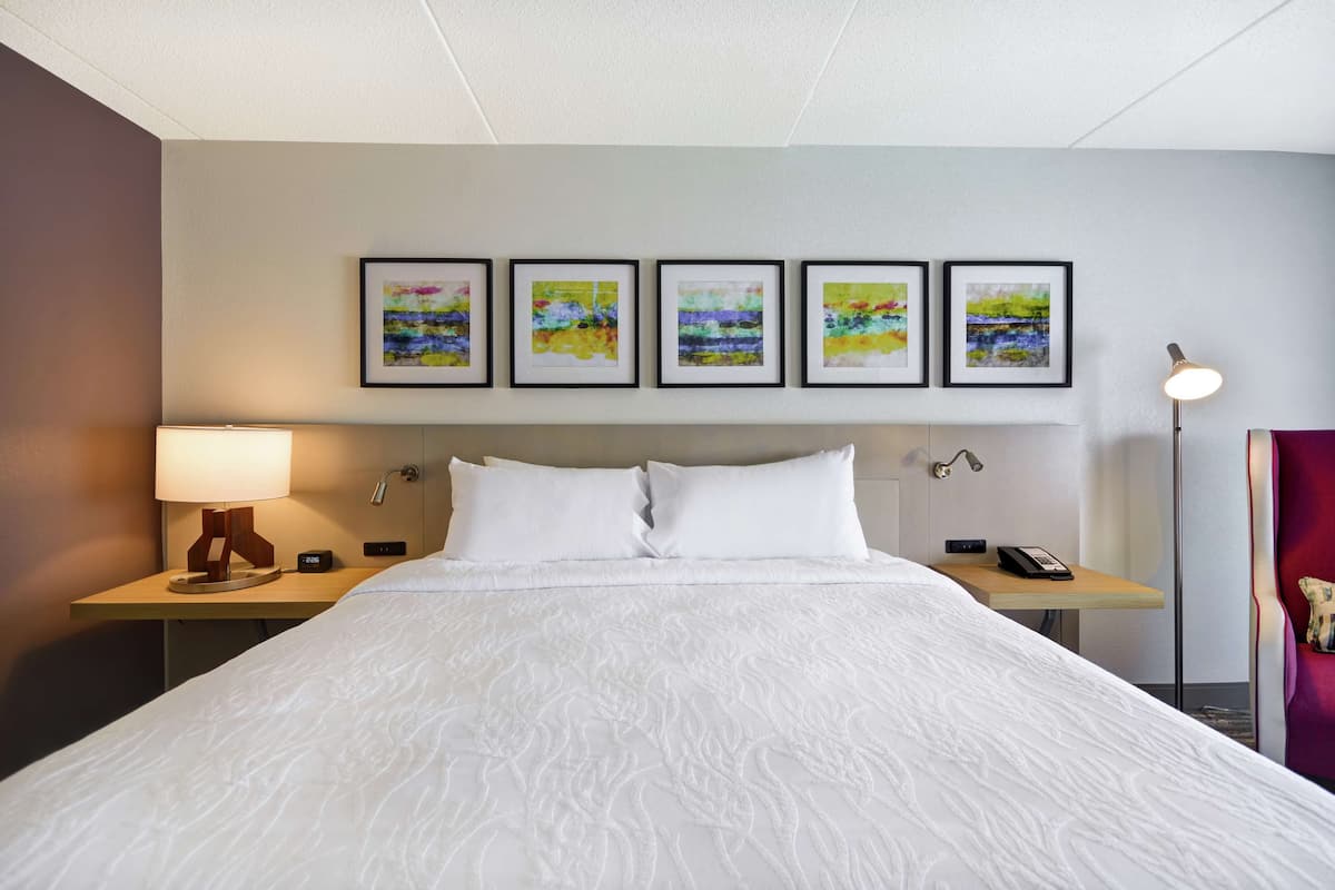 Une chambre d’hôtel avec un grand lit, une table de chevet avec une lampe, un téléphone et des œuvres d’art encadrées accrochées au mur.