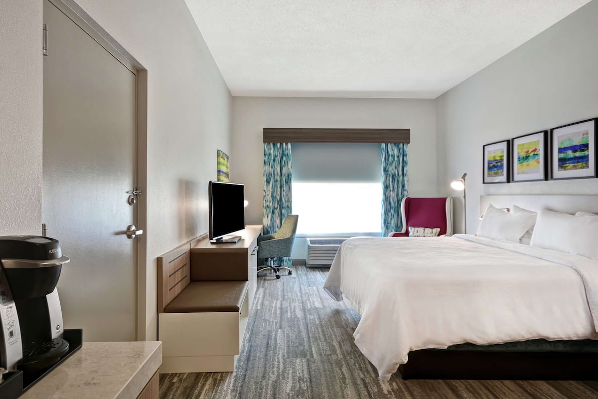 Une chambre d’hôtel comprenant un lit, un bureau avec un ordinateur, une chaise, une machine à café et une fenêtre avec des rideaux.