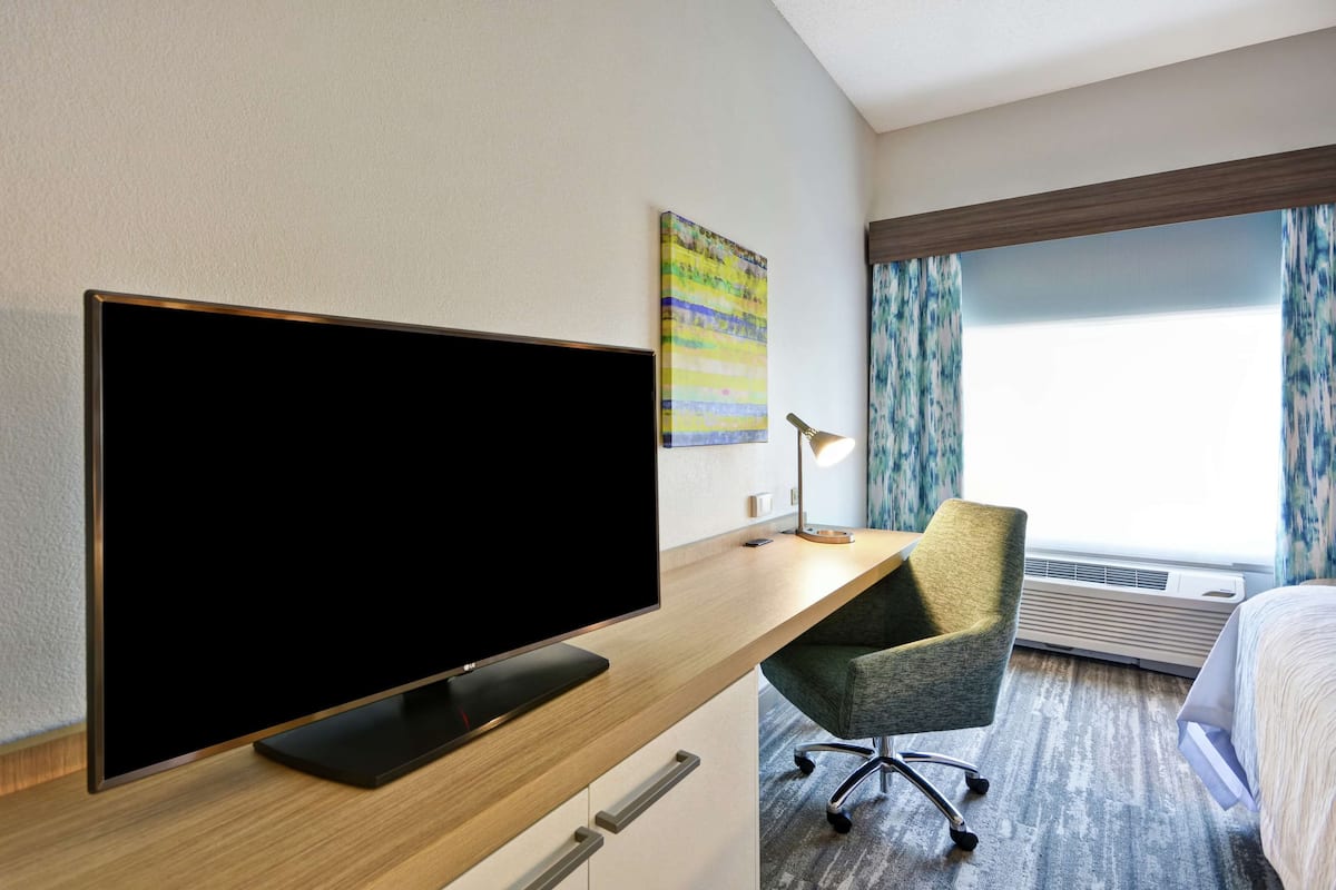 Une chambre d’hôtel équipée d’un grand téléviseur à écran plat, d’un bureau avec une chaise, d’un lit, d’une fenêtre avec des rideaux et d’un appareil de climatisation.