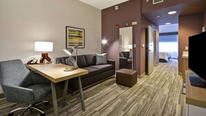 Ein Hotelzimmer mit Schreibtisch, Stuhl, Sofa und Hocker. Ein Fernseher, eine Lampe und ein Gemälde sind an der Wand zu sehen.