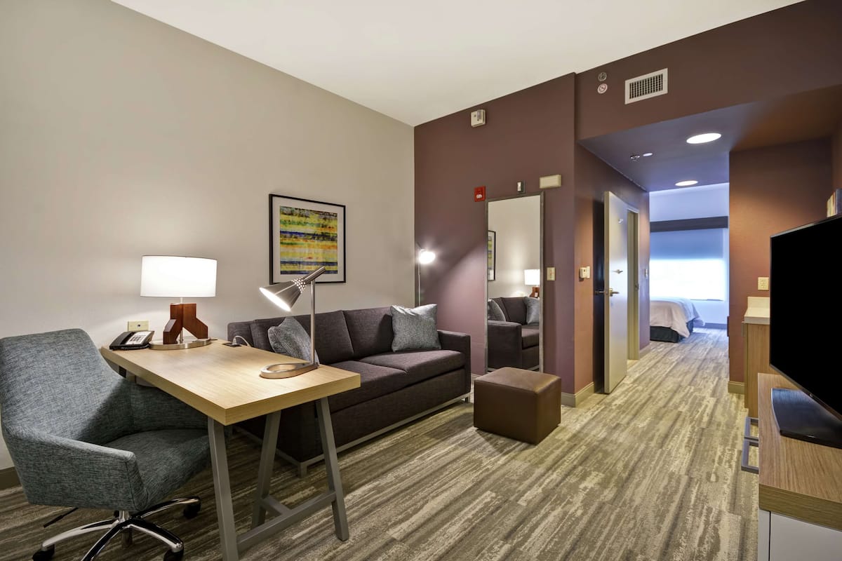 Une chambre d’hôtel équipée d’un bureau, d’une chaise, d’un canapé et d’un pouf. On y trouve également une télévision, une lampe et un tableau au mur.