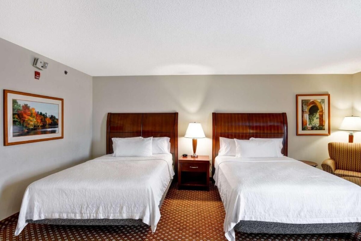 Una habitación de hotel con dos camas, una mesita de noche con lámpara, una silla y cuadros en las paredes.
