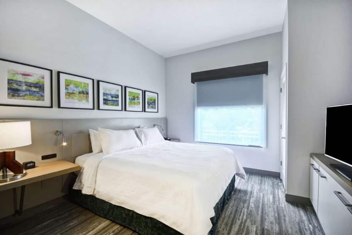 Une chambre d’hôtel avec un lit, une table de chevet, une lampe, un tableau au mur et une fenêtre avec des stores.