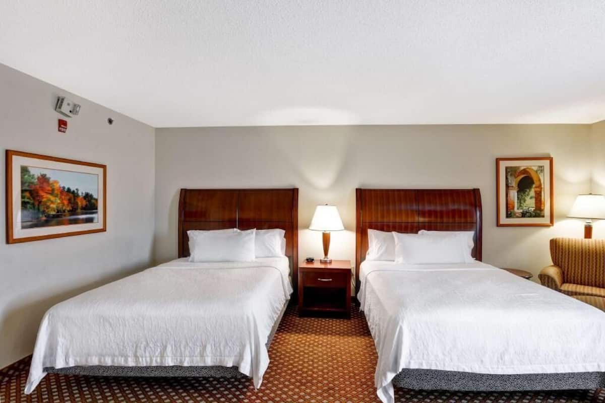Une chambre d’hôtel avec deux lits, une table de chevet avec une lampe, une chaise et des tableaux encadrés aux murs.