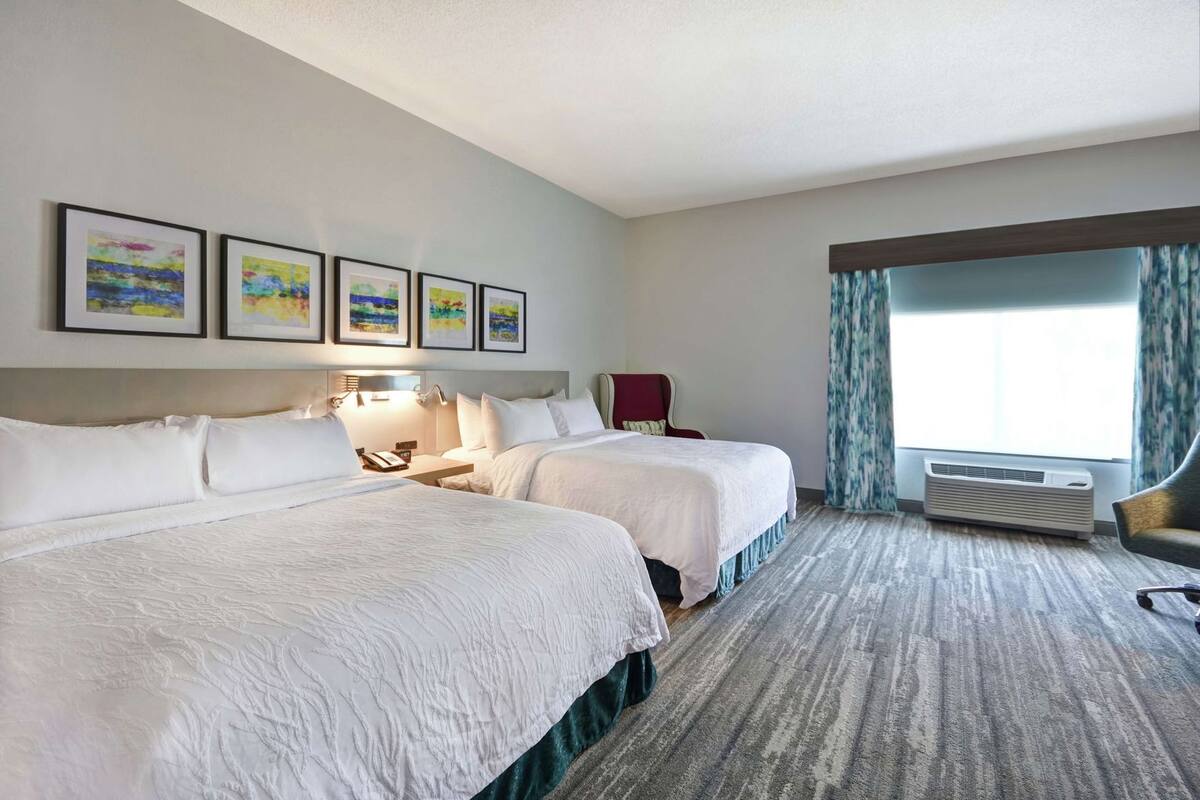 Une chambre d’hôtel avec deux lits, un bureau, une chaise, une fenêtre avec des rideaux et des œuvres d’art encadrées accrochées au mur.