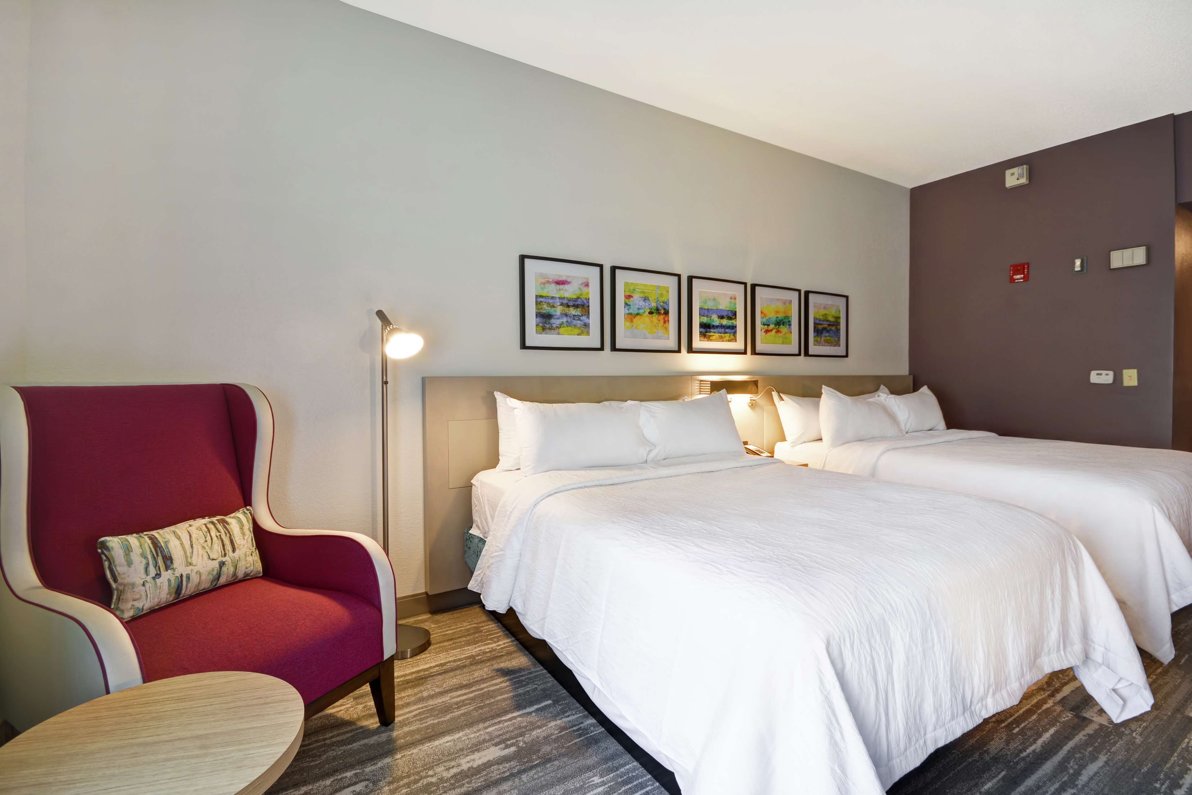 A hotel room with a red armchair, two beds with white bedding, a round table, and four framed artworks on the wall.