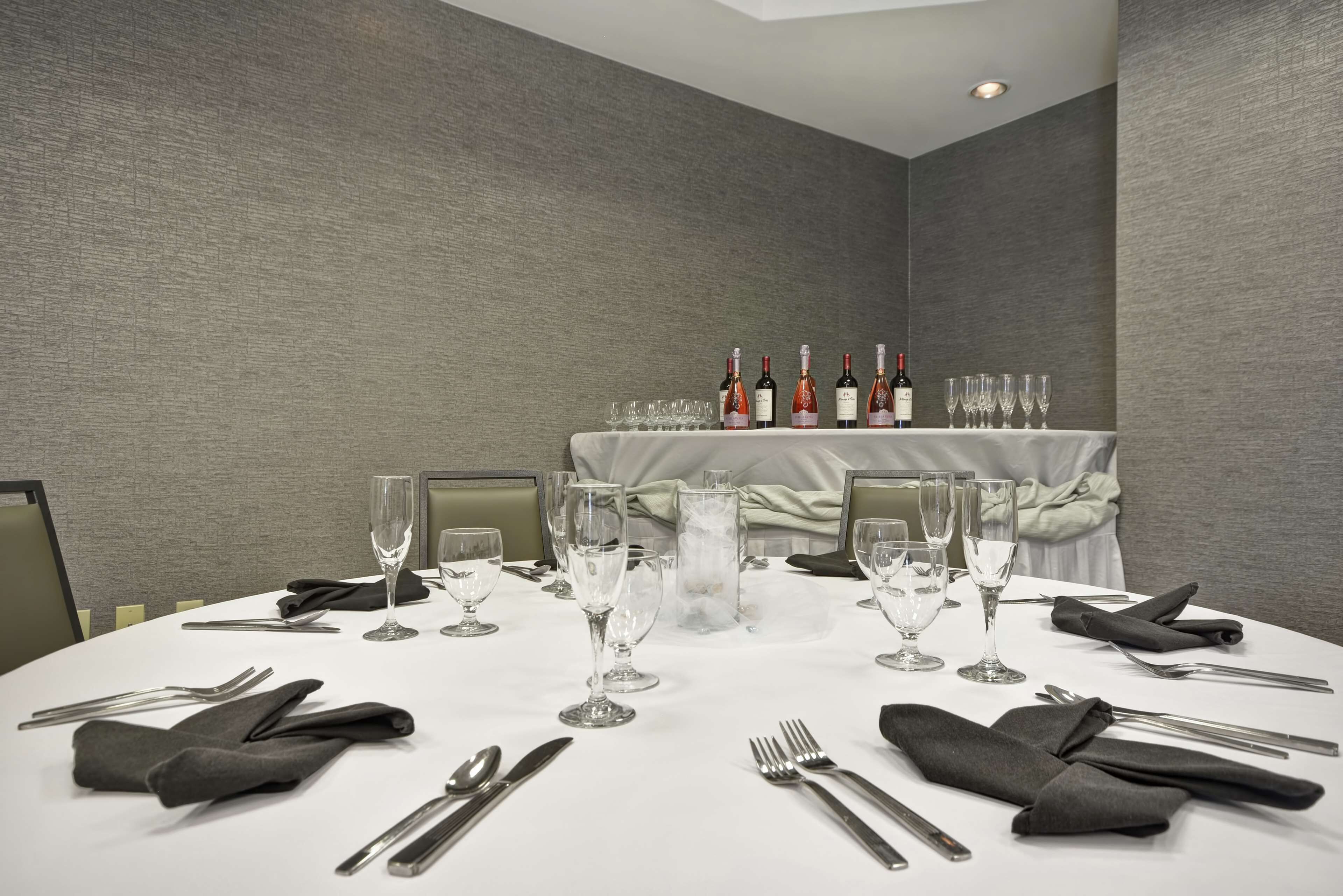 A round dining table set with wine glasses, bottles, and cutlery, with a backdrop of grey walls and a tablecloth-covered table.