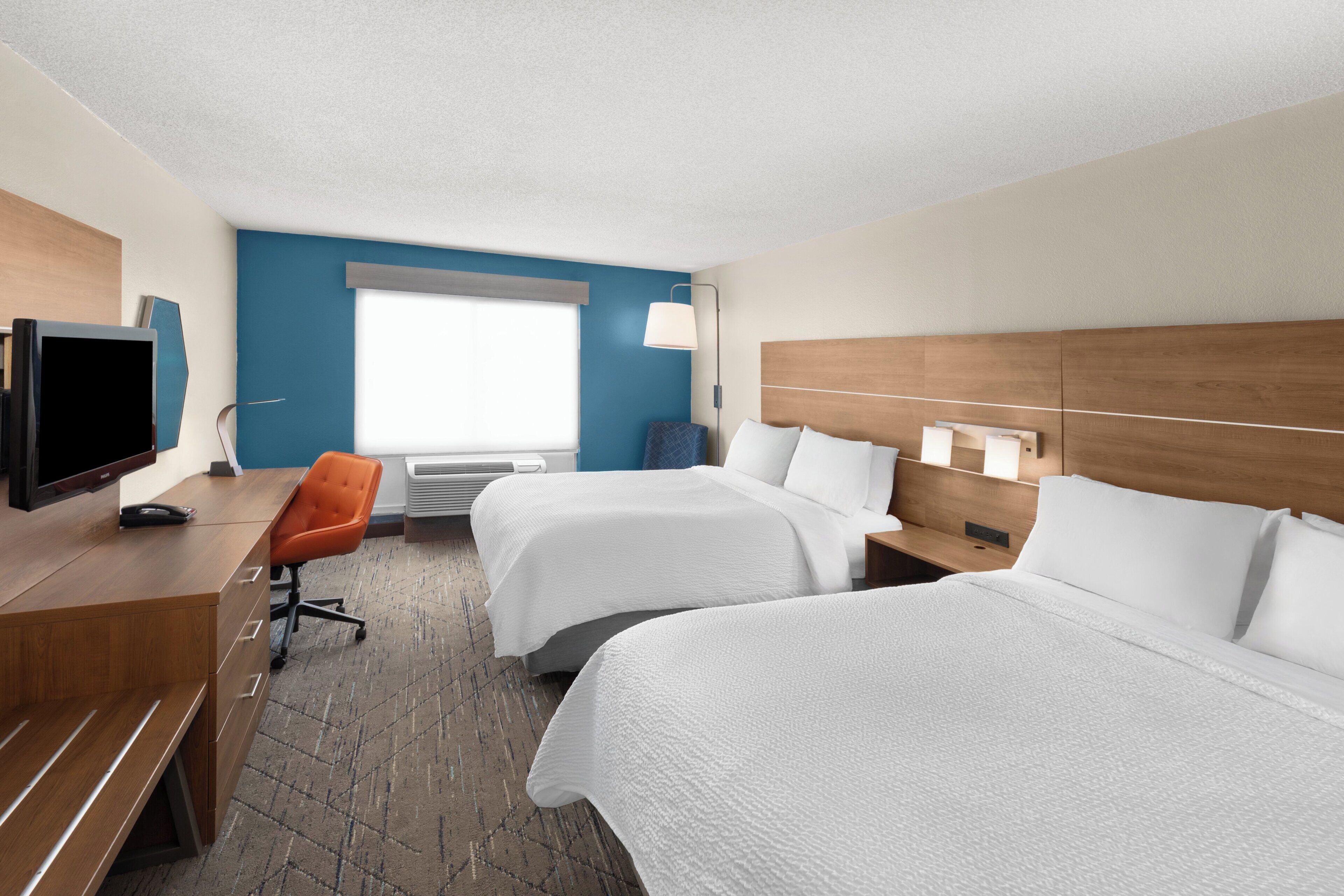 A hotel room with two beds, a desk with a TV, a chair, a window, and a lamp.
