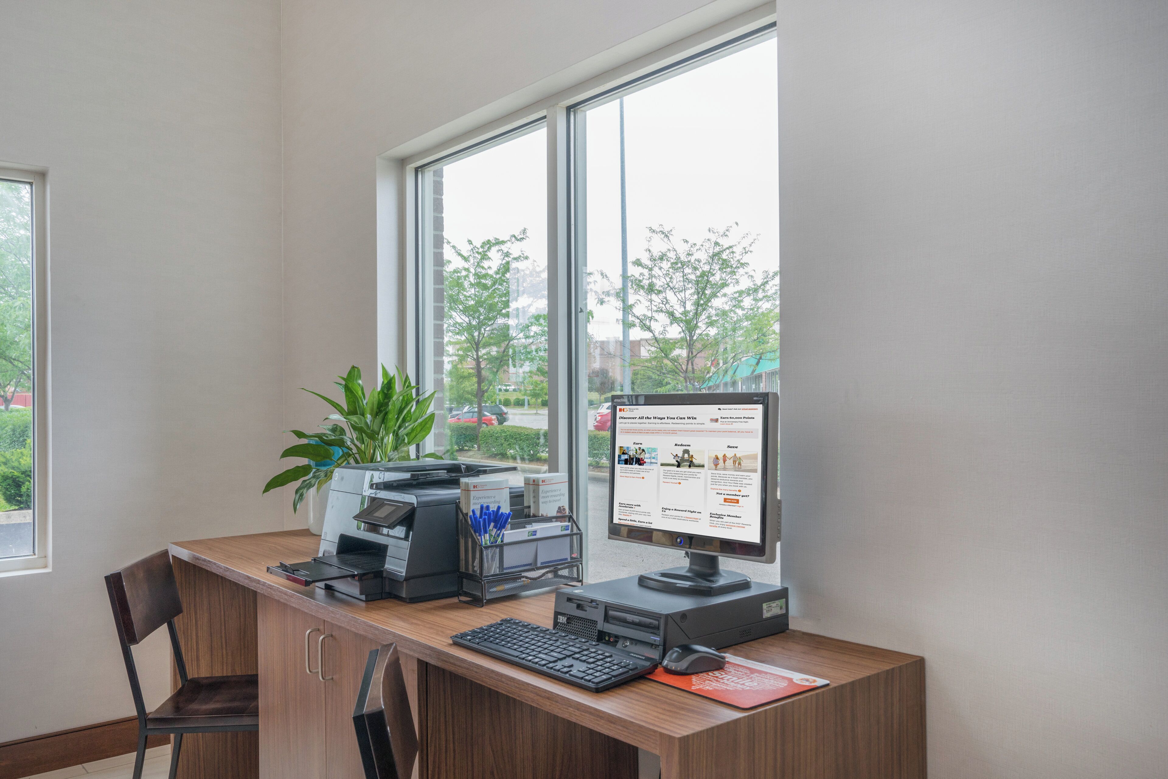 Escritorio con computadora, impresora y teclado, ubicado cerca de una ventana que ofrece vistas a árboles y edificios.