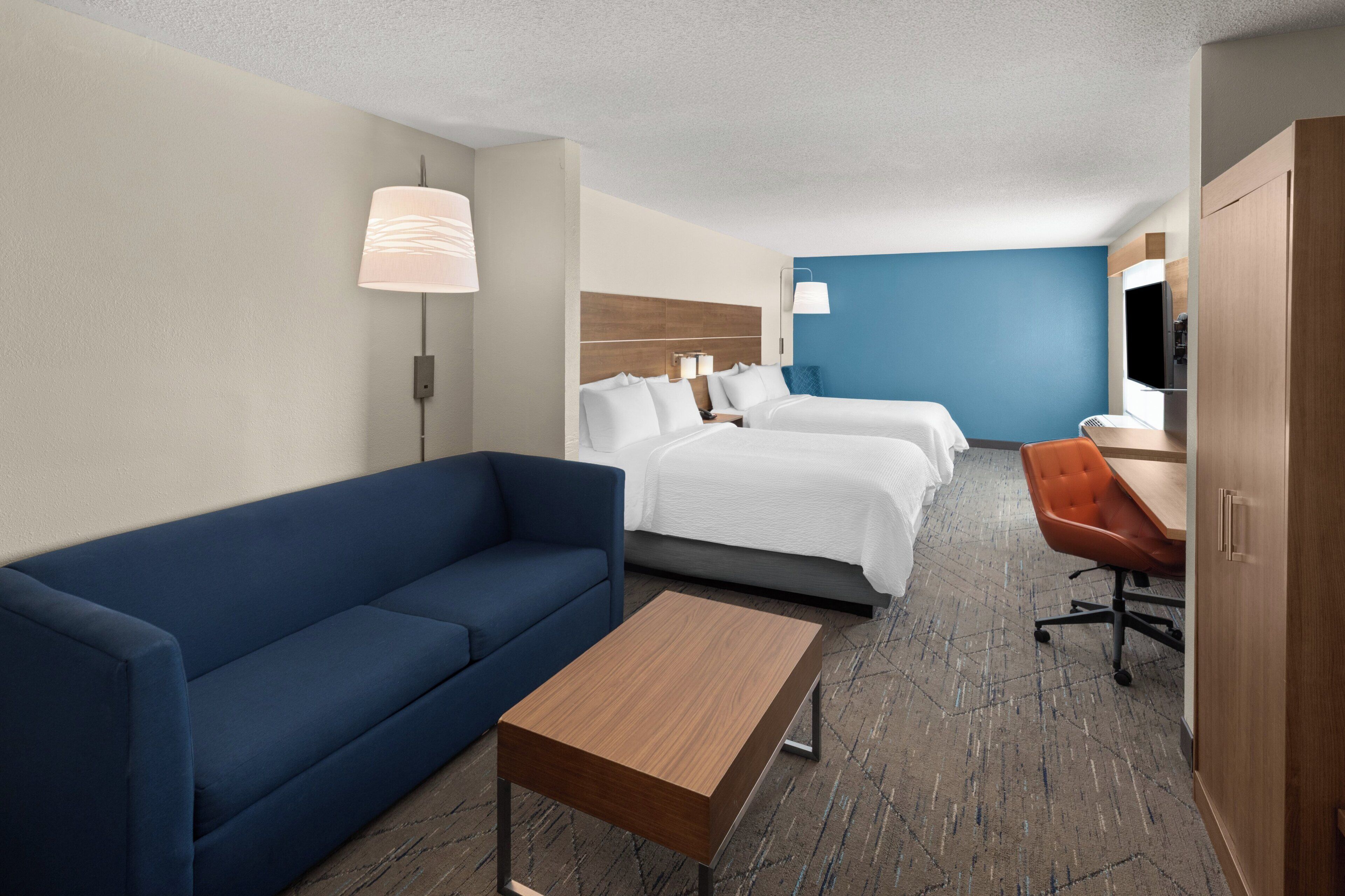 Habitación de hotel con un sofá azul, dos camas individuales, una mesa de madera, un escritorio con una silla y un televisor.