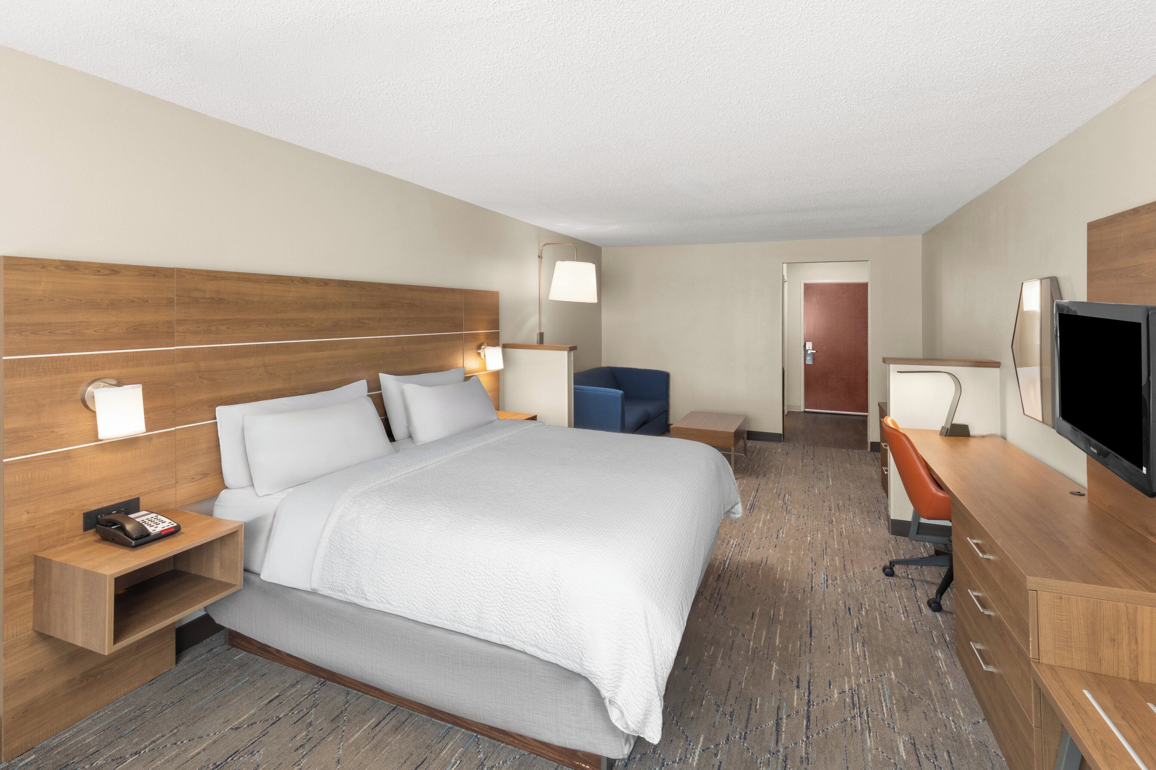 A hotel room with a large bed, bedside tables, a desk with a chair, a television, and a door leading to another room.