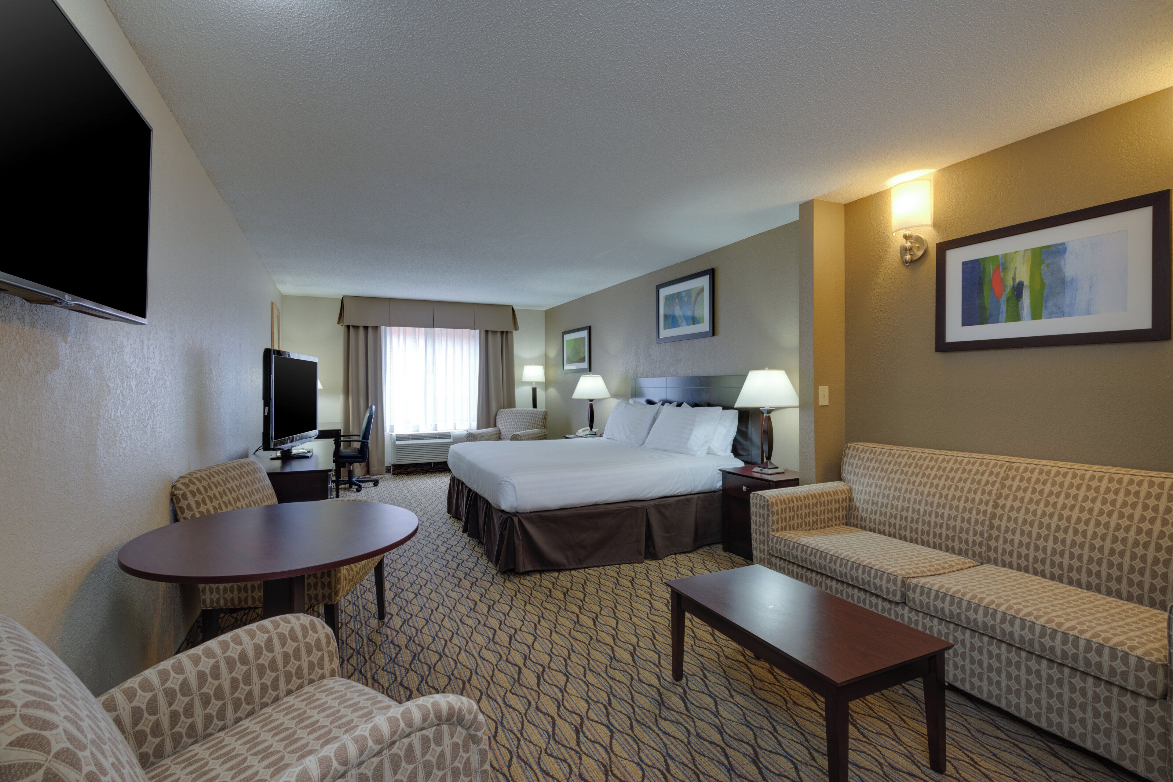 Une chambre d’hôtel comprenant un lit, deux chaises, un canapé, une petite table, un téléviseur et un bureau avec une lampe.