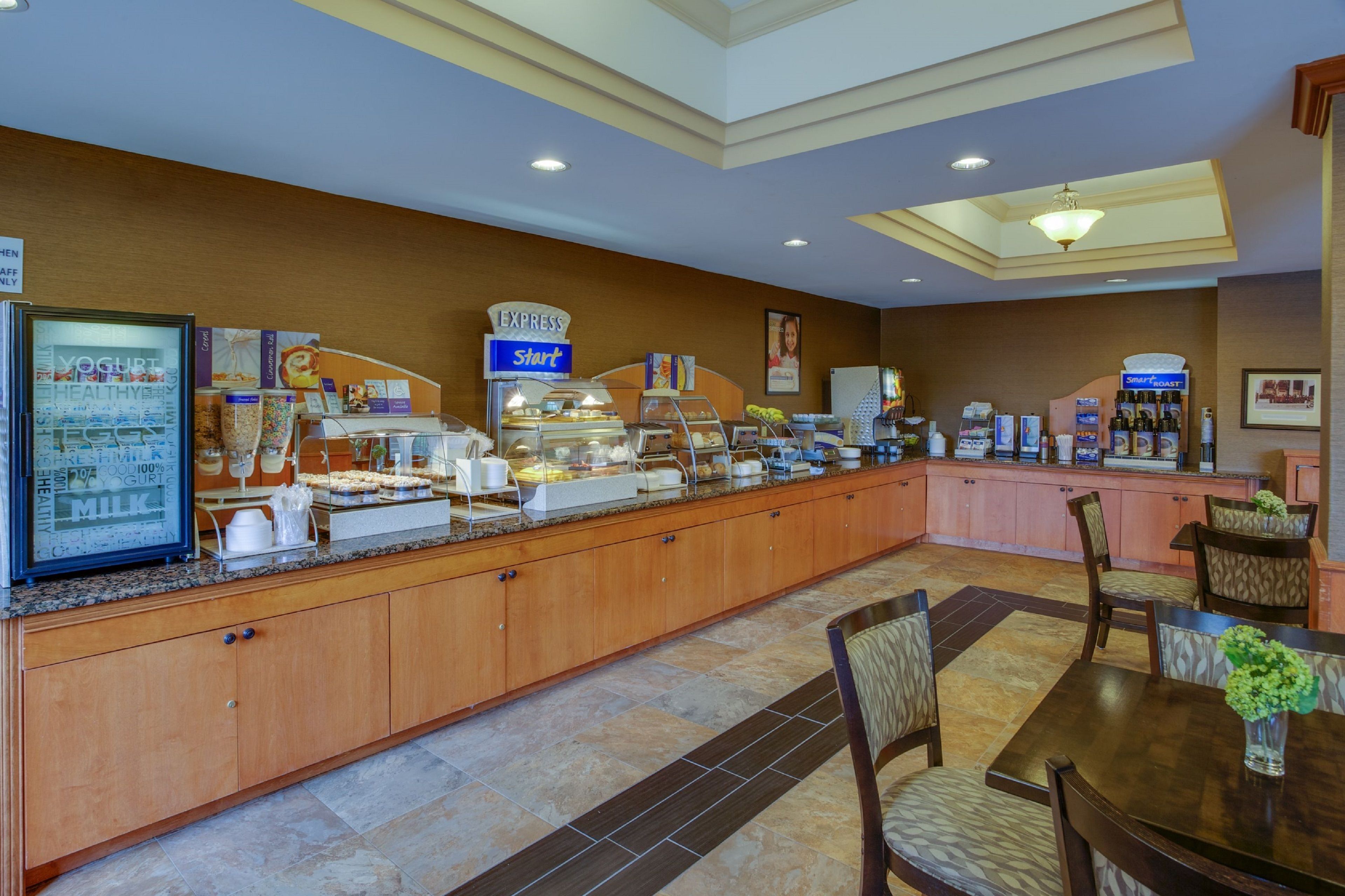 Uma área de buffet com diversos pontos de serviço de comida, incluindo uma seleção de iogurtes e uma estação de café. Há armários de madeira e piso cerâmico.