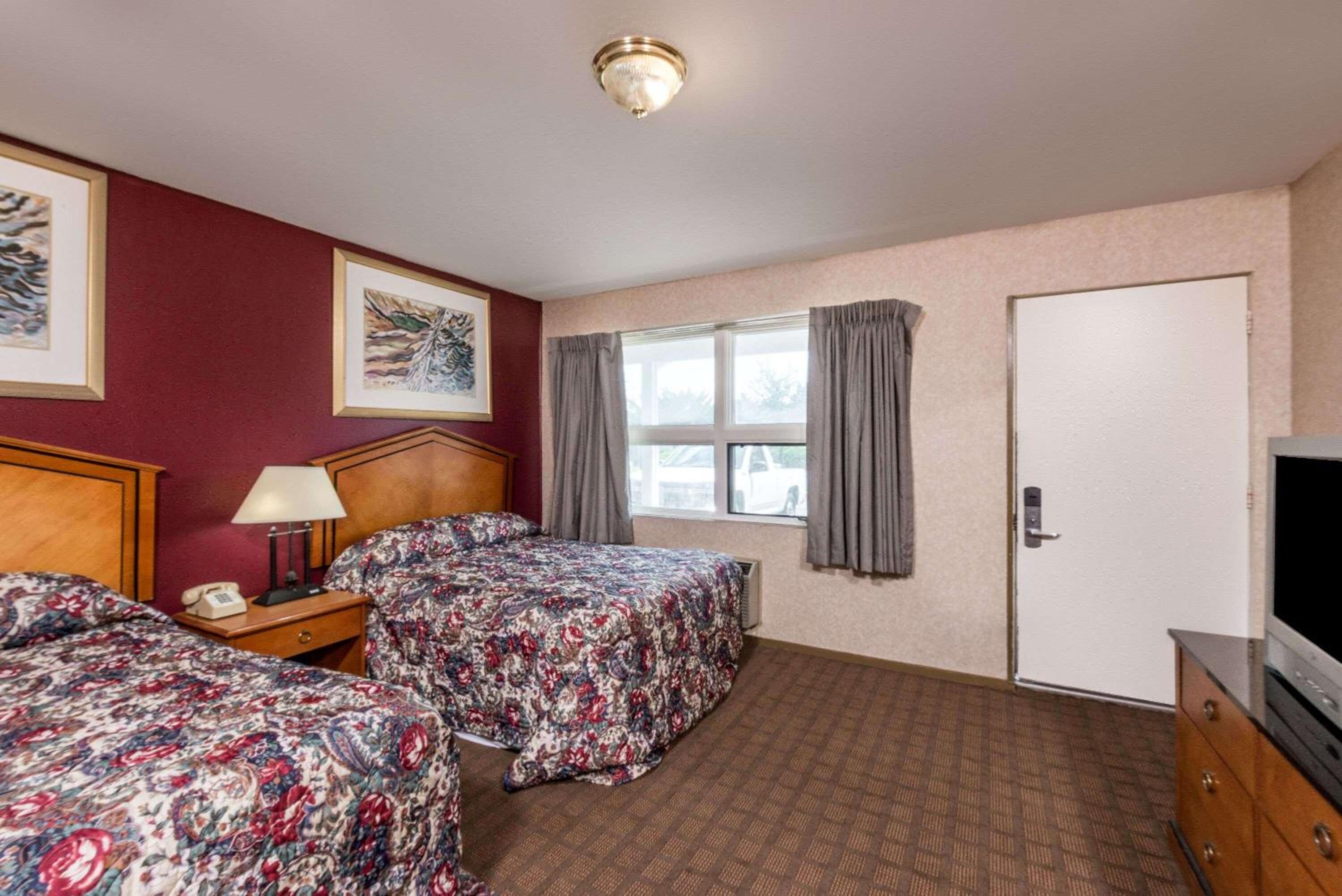 Habitación de hotel con dos camas, televisión, escritorio con lámpara, puerta y ventana con cortinas.