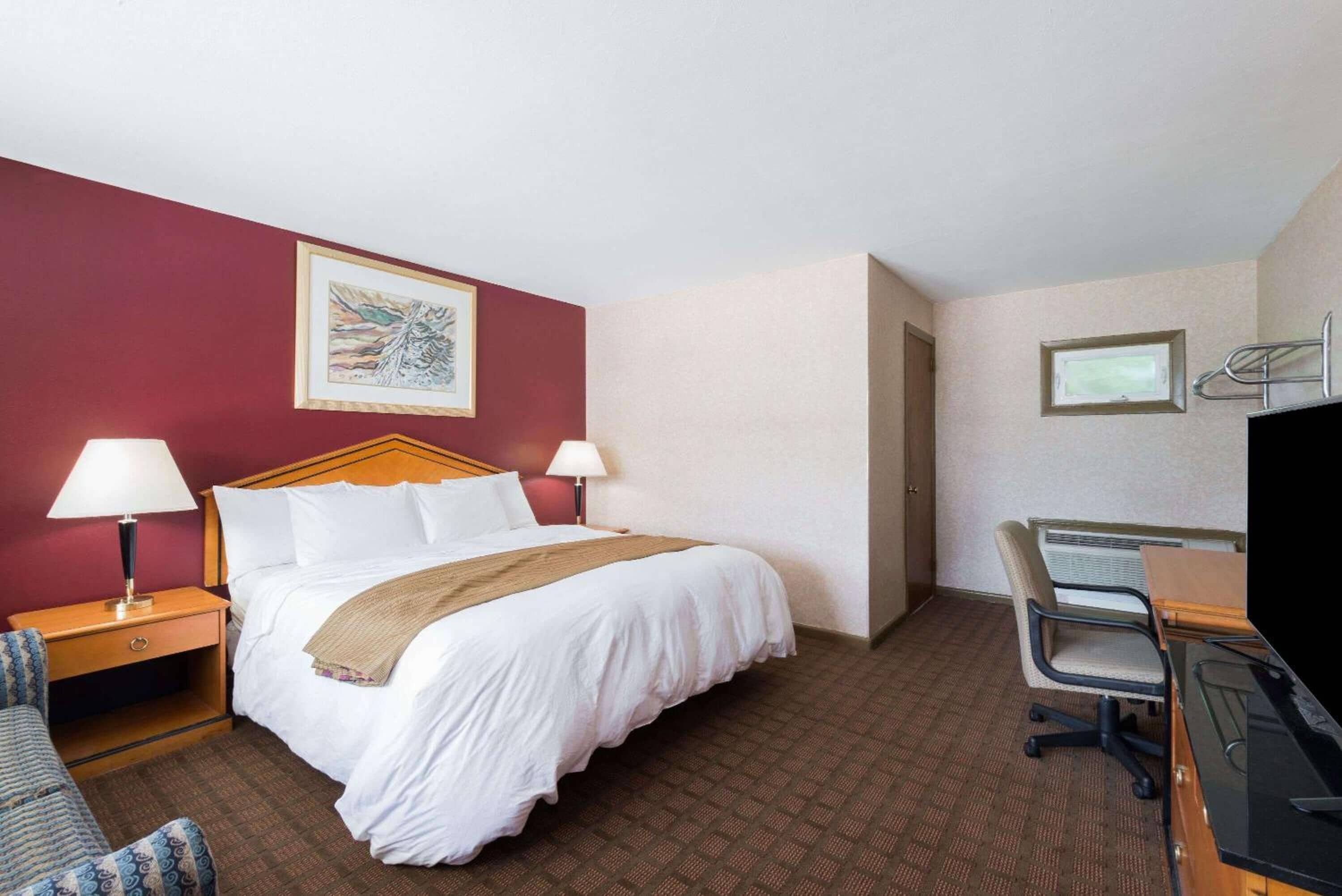 A hotel room with a bed, two bedside lamps, a desk with a chair, a TV, and a framed picture on the wall.