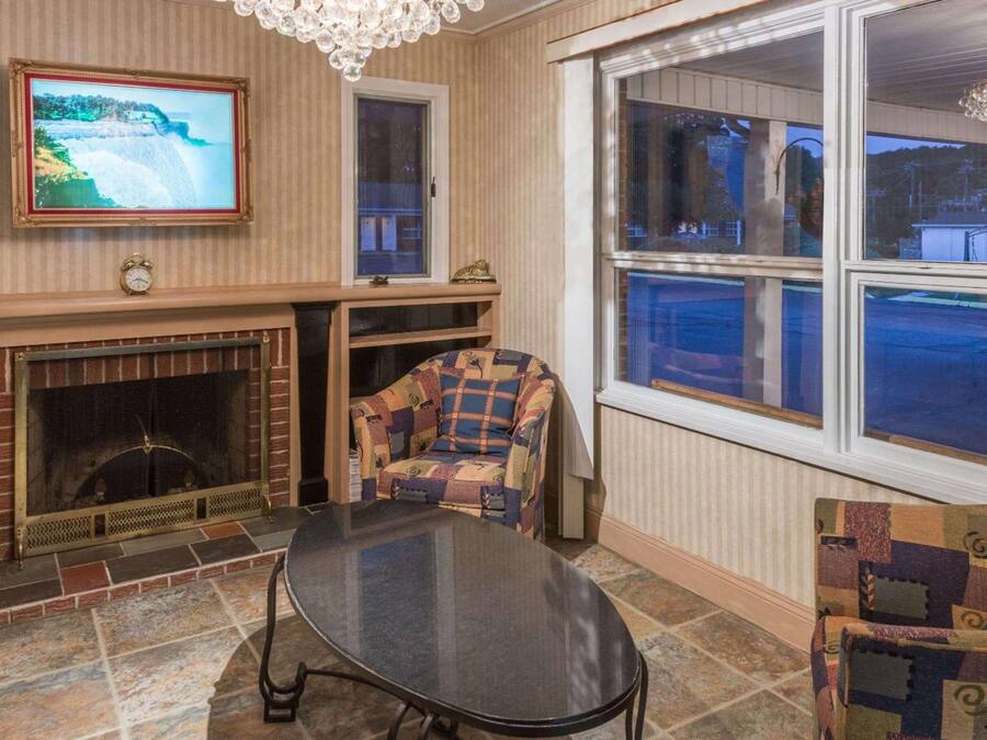 A living room with a fireplace, a television mounted on the wall, a patterned armchair, and a large window with a view of the outside.