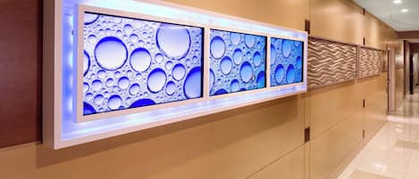 Um corredor com vários ecrãs fixados na parede, exibindo padrões azuis abstratos, uma parede texturizada ondulada e chão em mosaico.