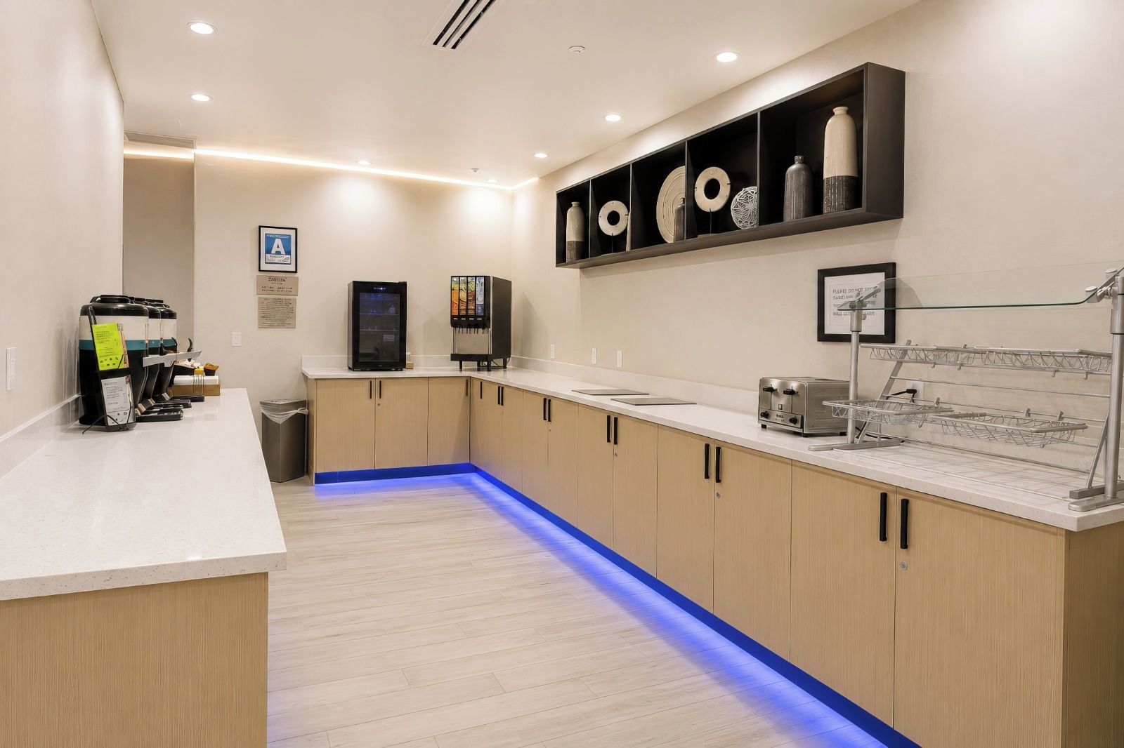 A modern kitchen with a white countertop, wooden cabinets, and a blue-lit floor. There are coffee machines, a refrigerator, and a toaster on the counter.