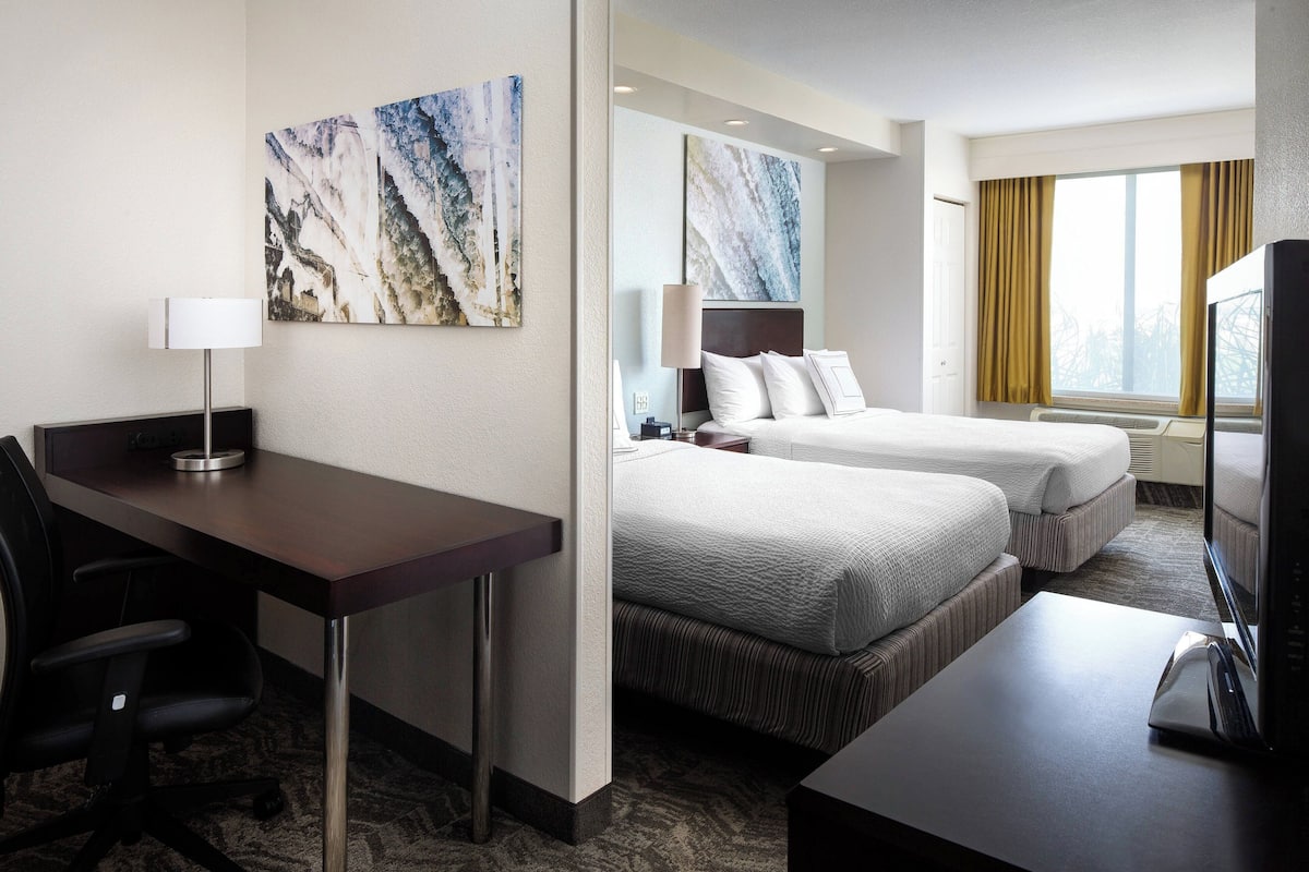 A hotel room with a desk, two beds, a TV, and artwork on the walls.