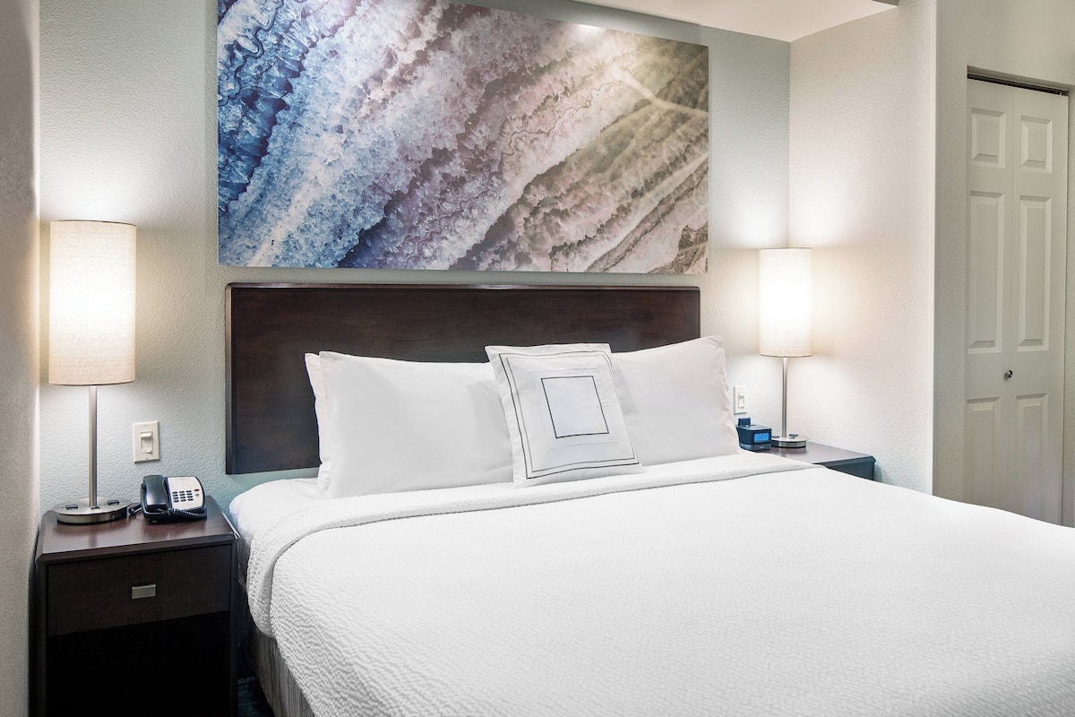 A hotel room with a large bed, two bedside lamps, a nightstand with a phone, and a wall-mounted artwork.