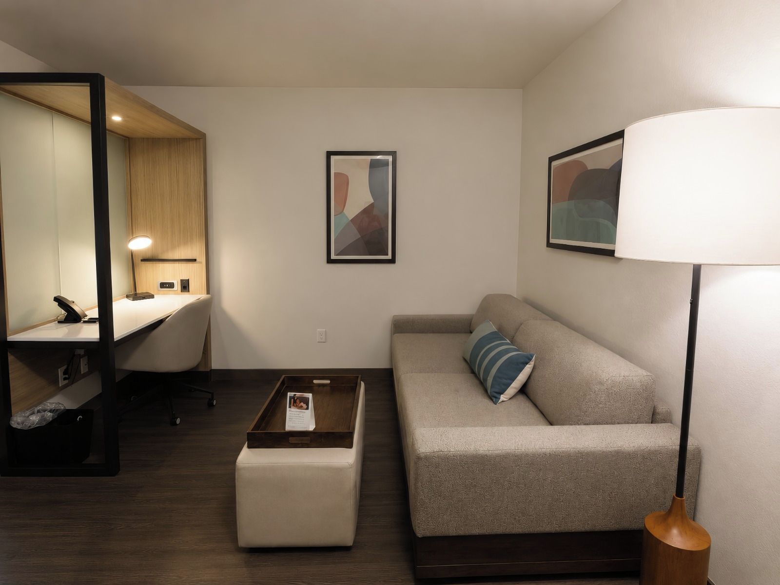 A modern hotel room with a desk, a sofa, a coffee table, a lamp, and artwork on the walls.